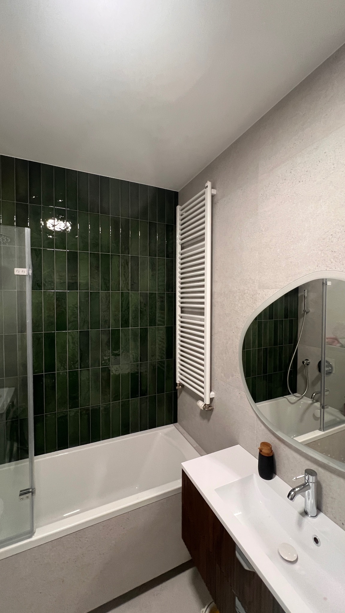 Wykończona łazienka z wanną obłożoną pionowymi, ciemnozielonymi płytkami, grzejnikiem drabinkowym i owalnym lustrem nad umywalką z szafką w odcieniach brązu.
