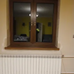 Zamknięte, dwuskrzydłowe okno z brązową ramą, widoczne na tle ściany w odcieniach beżu i grzejnika. W szybach odbija się wnętrze pokoju z żółtymi ścianami.