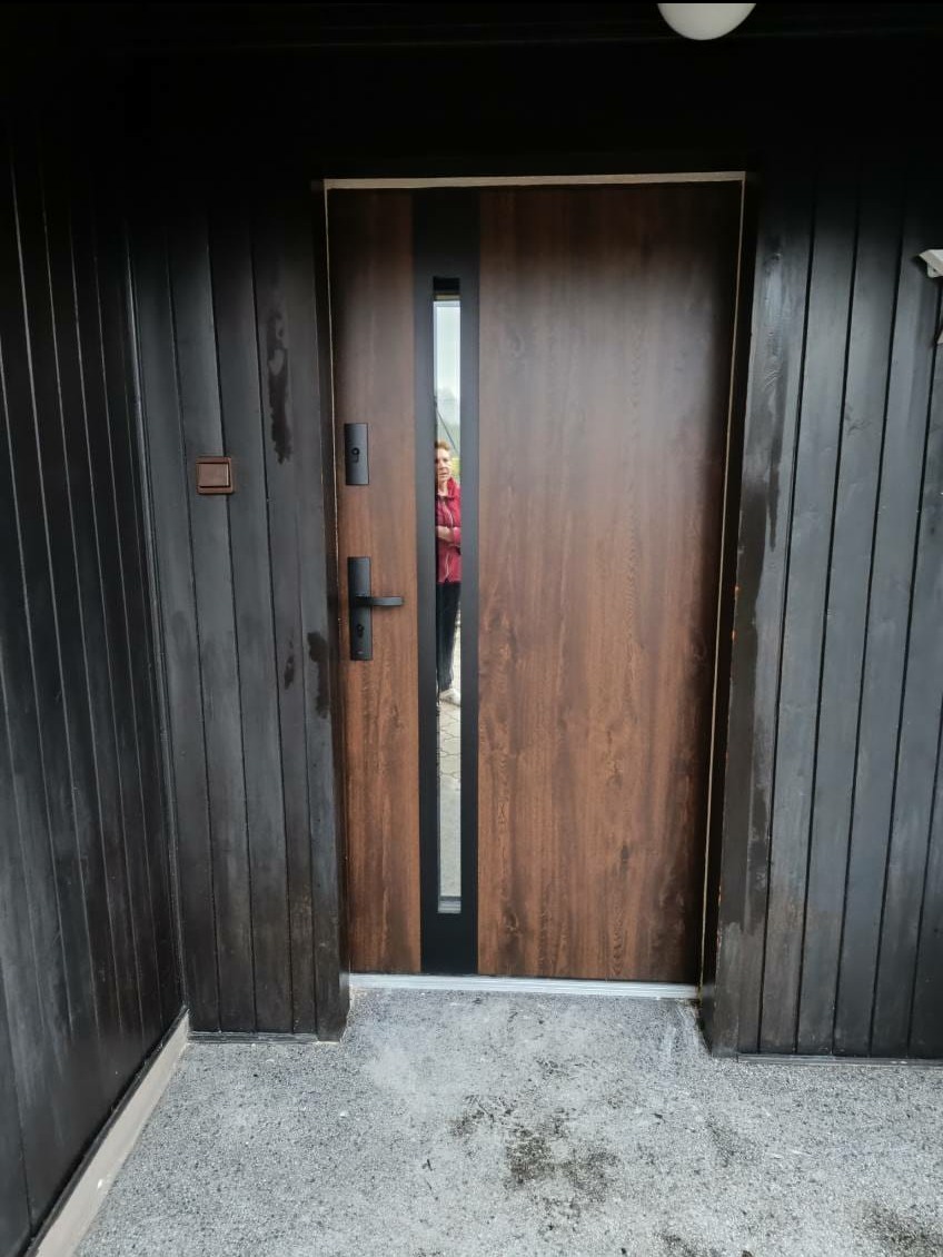 Brązowe drzwi wejściowe z czarnym panelem i klamką, zamontowane w ciemnym, drewnianym budynku. W szybie drzwi odbija się sylwetka osoby.