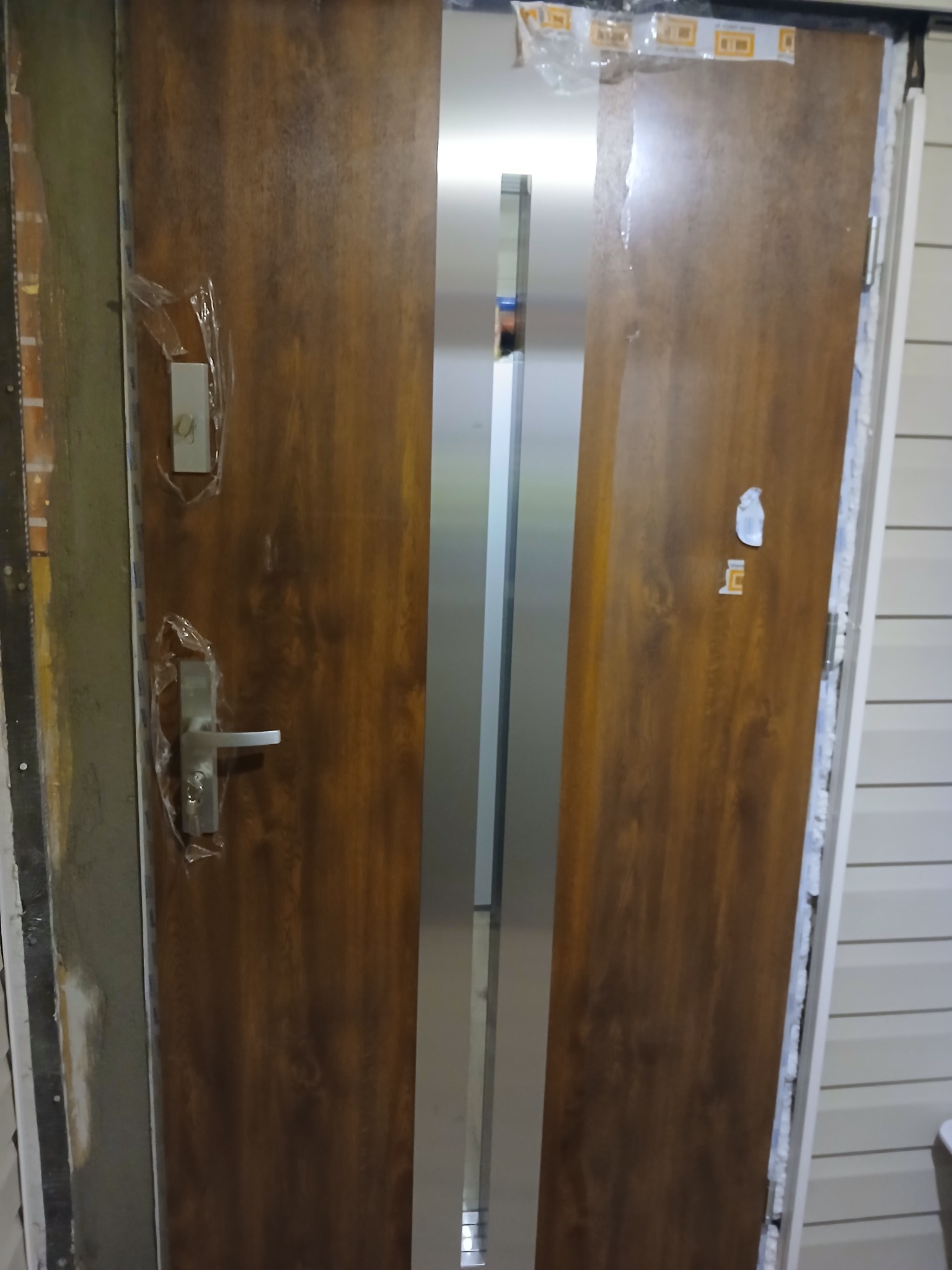 Nowe, brązowe drzwi wejściowe z srebrnym pasem pośrodku, zabezpieczone folią ochronną, w trakcie montażu, widoczne pianka montażowa i niedokończone wykończenie.