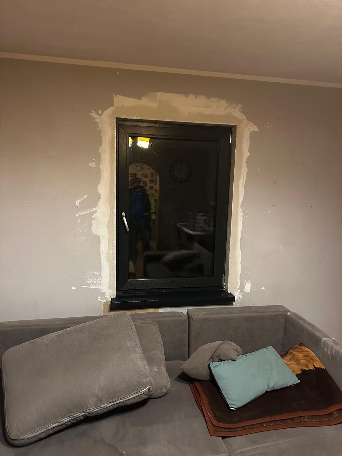 Czarne okno po montażu w szarej ścianie z widocznymi śladami gładzi, odblask z sylwetką osoby w szybie, fragment sofy z poduszkami na pierwszym planie.