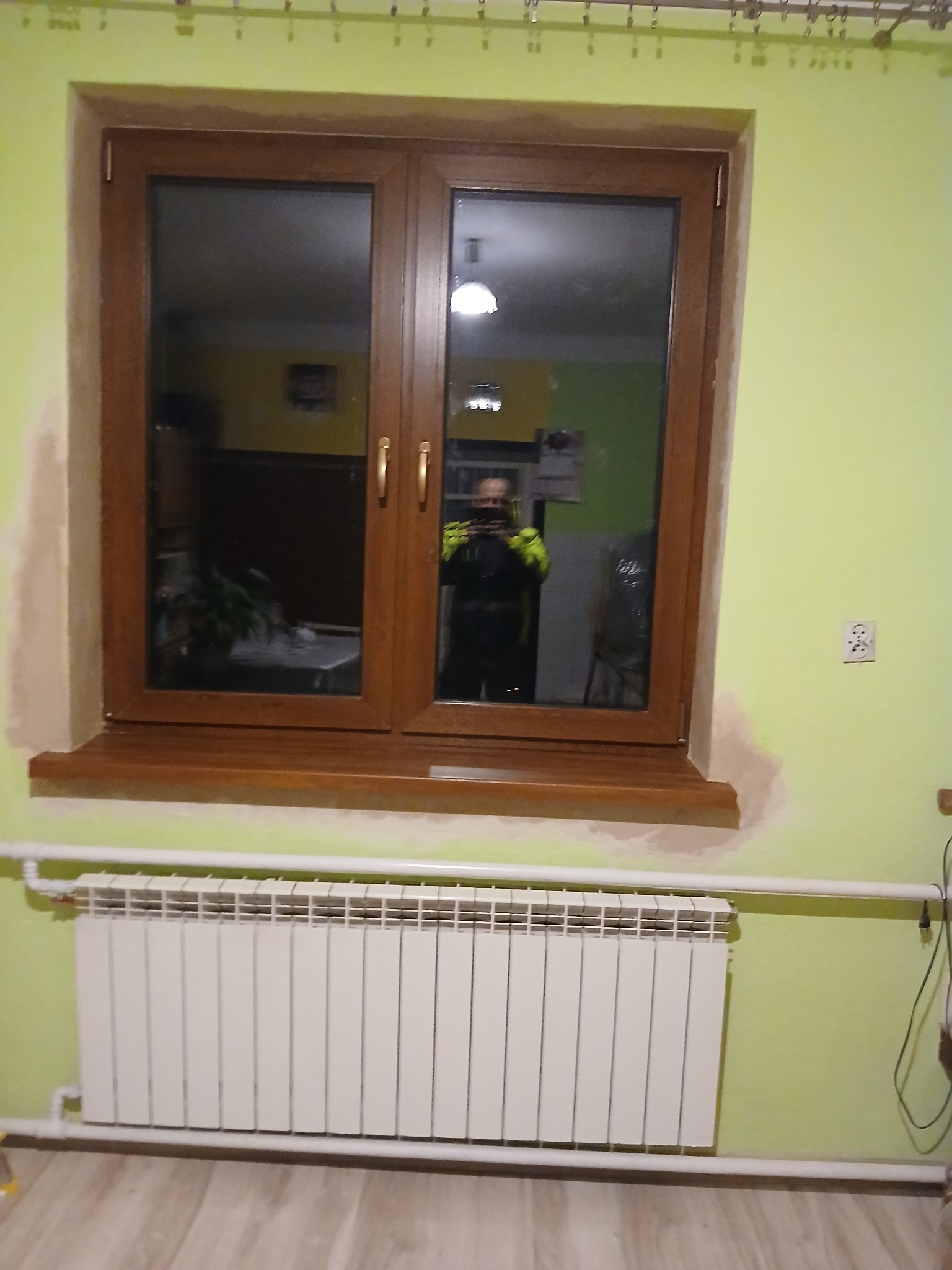 Brązowe okno PCV z widocznym odbiciem osoby w środku, zamontowane na tle jasnozielonej ściany z widocznym grzejnikiem i częściowo przygotowaną ścianą do malowania.