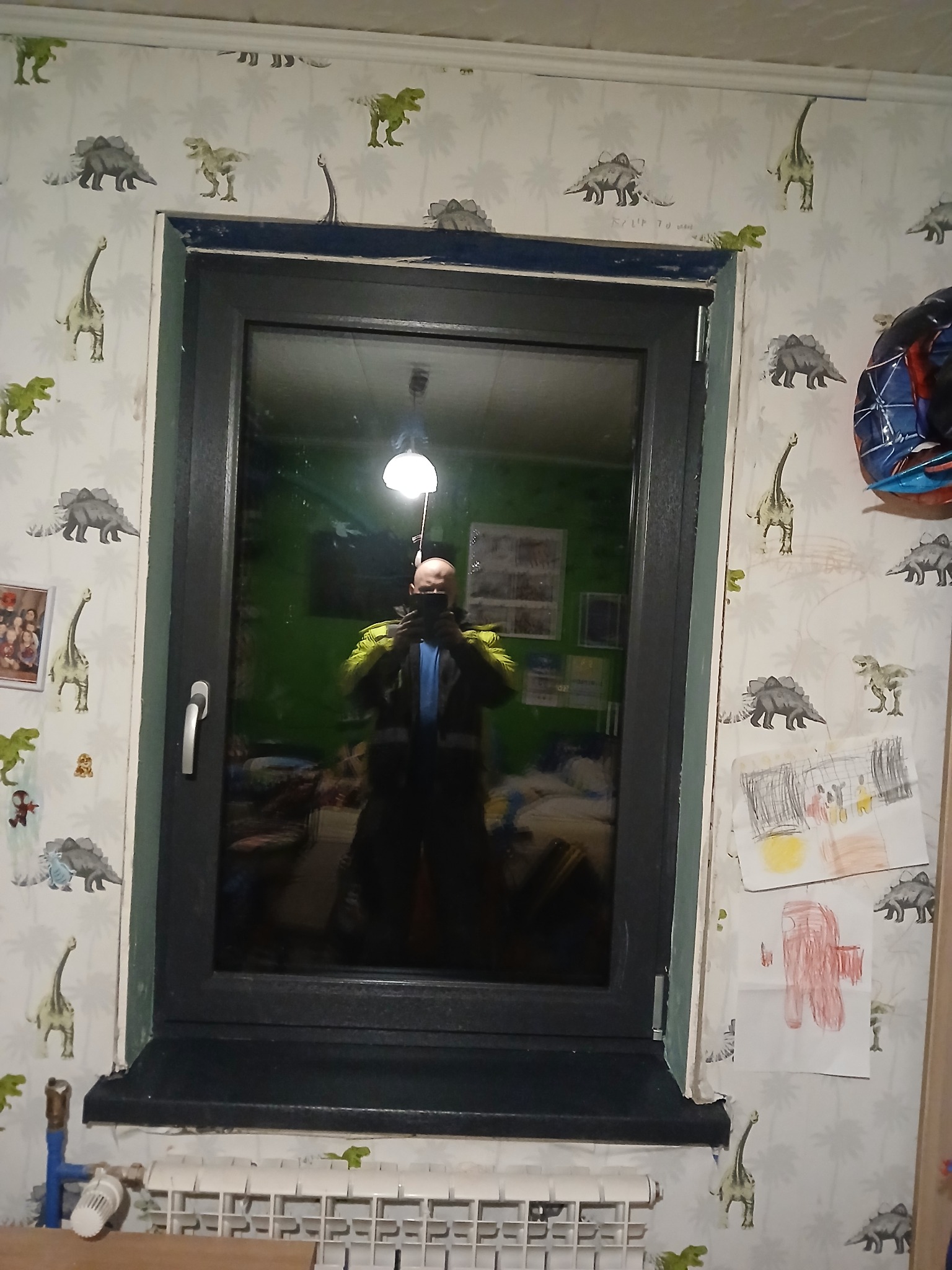 Szare okno PCV w pokoju dziecięcym z tapetą w dinozaury. W szybie odbija się osoba robiąca zdjęcie. Widoczny biały grzejnik pod oknem i rysunki na ścianie.