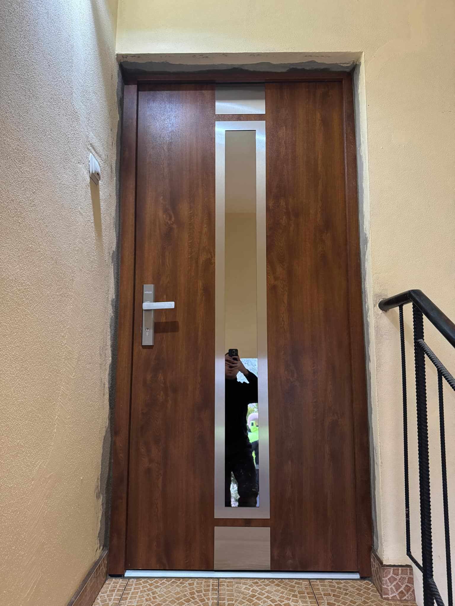 Zamontowane drewnopodobne drzwi wejściowe z szybą, widoczne na klatce schodowej. Świeży montaż, widoczne ślady zaprawy. Osoba w odbiciu szyby.