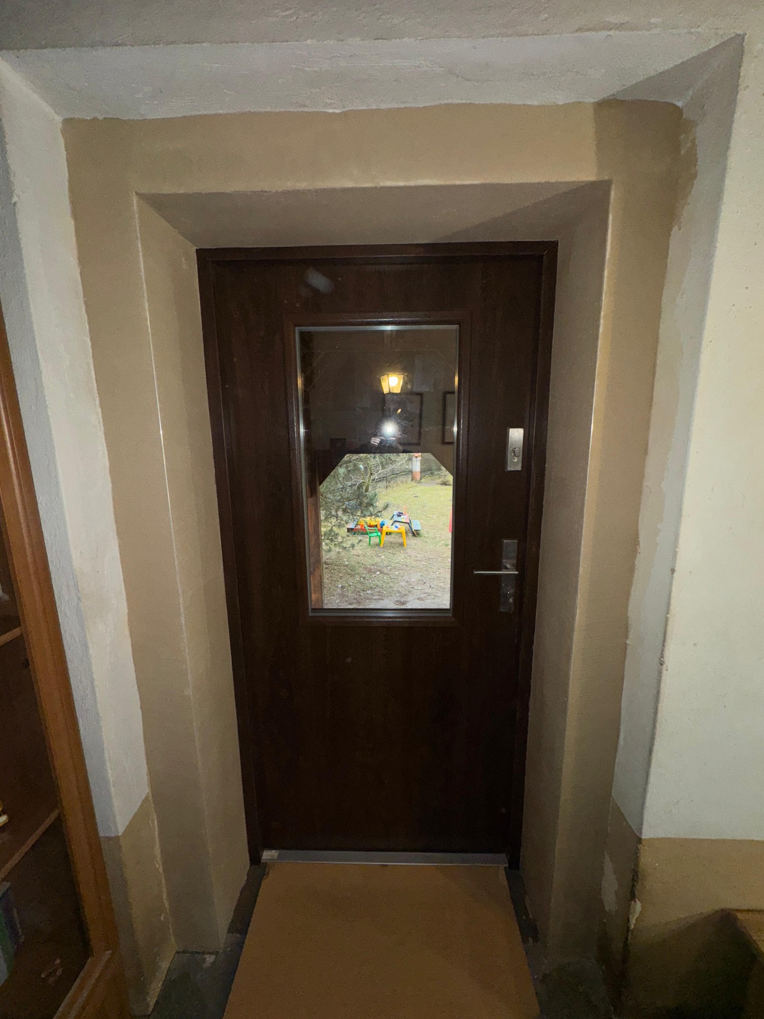 Ciemne, drewniane drzwi z szybą w beżowej futrynie. Przez szybę widoczny ogród z zabawkami. W tle rama obrazu i lampa.