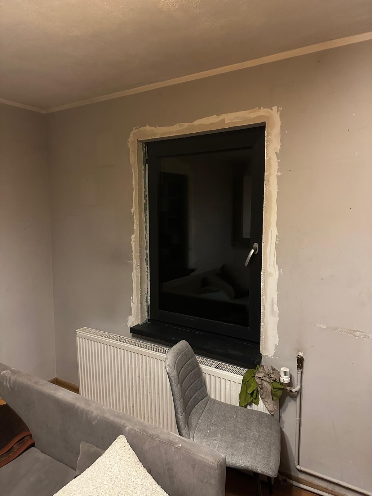 Czarne okno PCV po montażu, widoczne ślady po obróbce tynku. Wnętrze szarego pokoju z grzejnikiem i meblami. Ujęcie z bliska na okno.