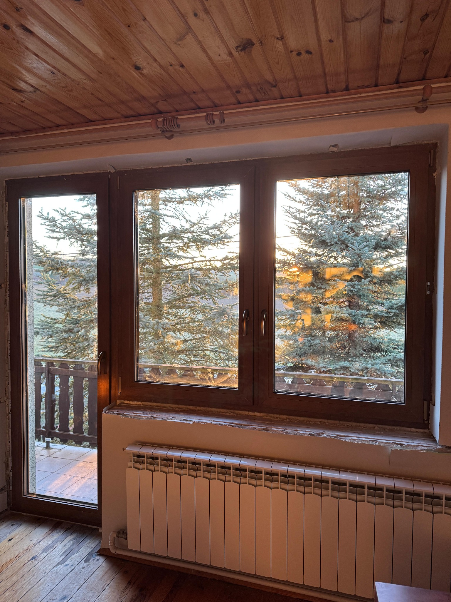 Trzyczęściowe okno z brązową ramą, widok na zewnątrz na balkon i drzewa iglaste w promieniach zachodzącego słońca. Drewniany sufit i grzejnik pod oknem.