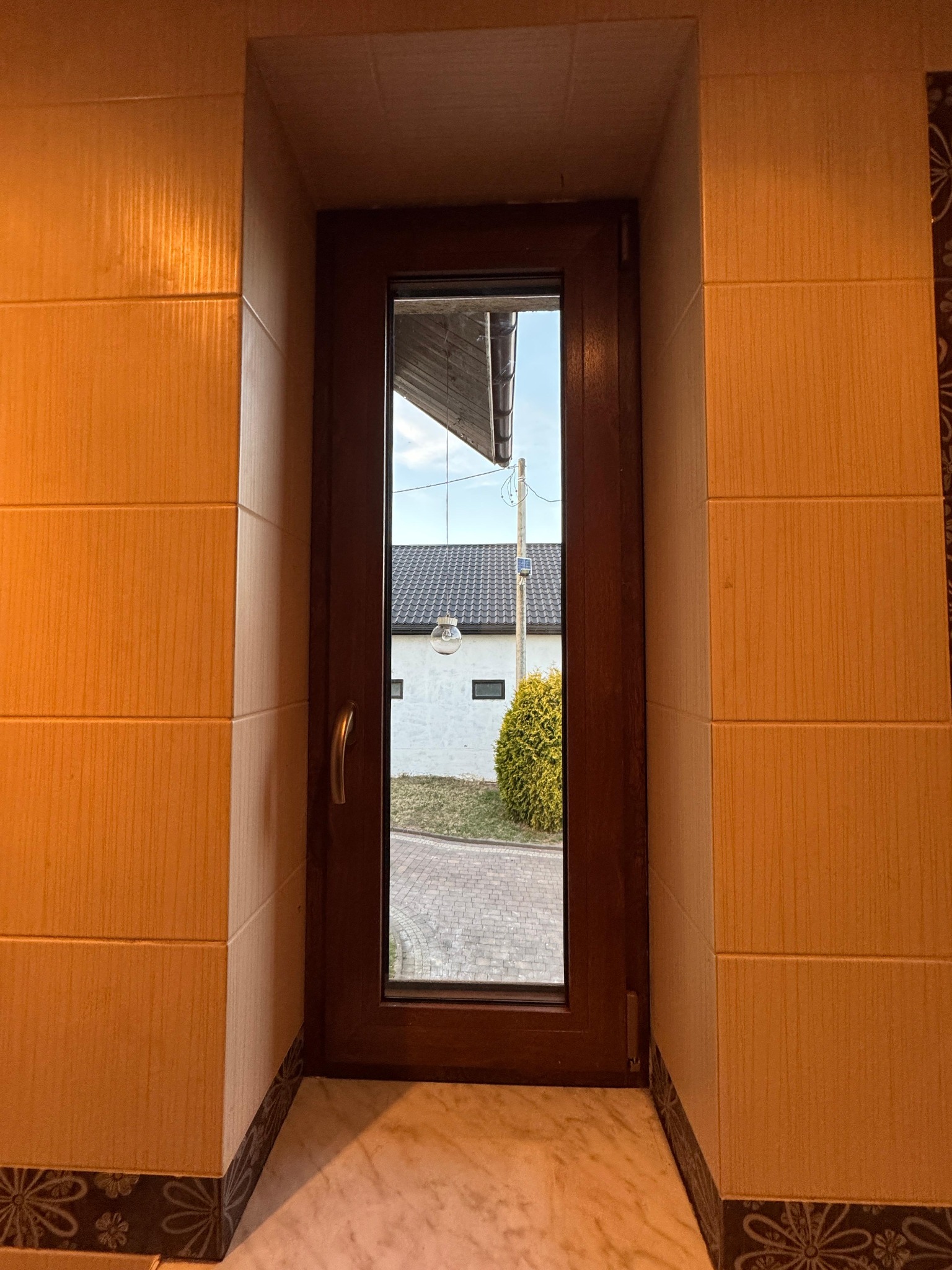 Wnętrze z oknem z brązową ramą, widok na ogród i dom. Okno osadzone w ścianie wyłożonej płytkami w ciepłych odcieniach beżu i brązu, marmurowy parapet.