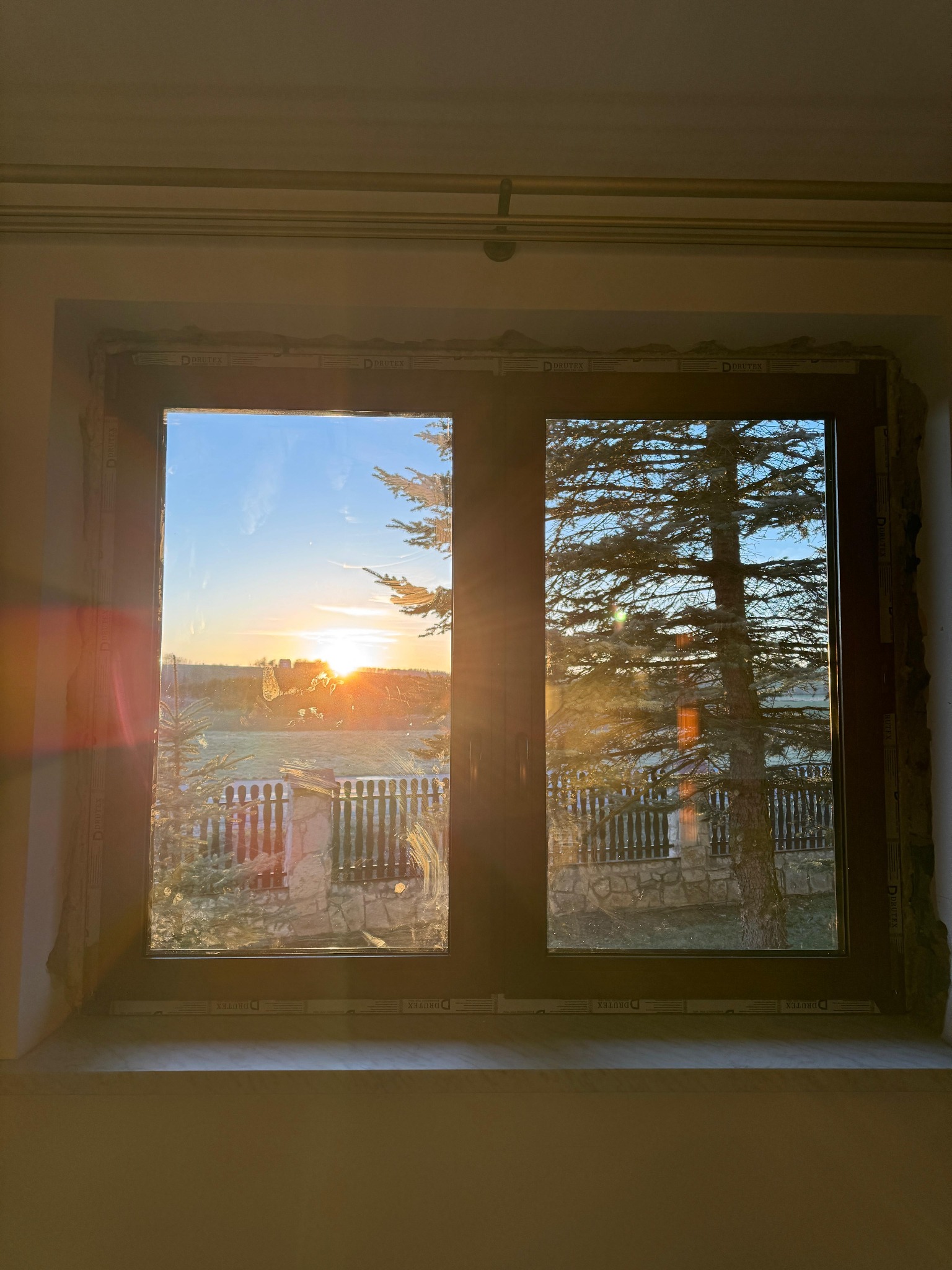 Nowe okno z widokiem na wschód słońca i drzewo iglaste. Profil okienny DRUTEX. Szyba lekko zabrudzona. W tle widoczny krajobraz.