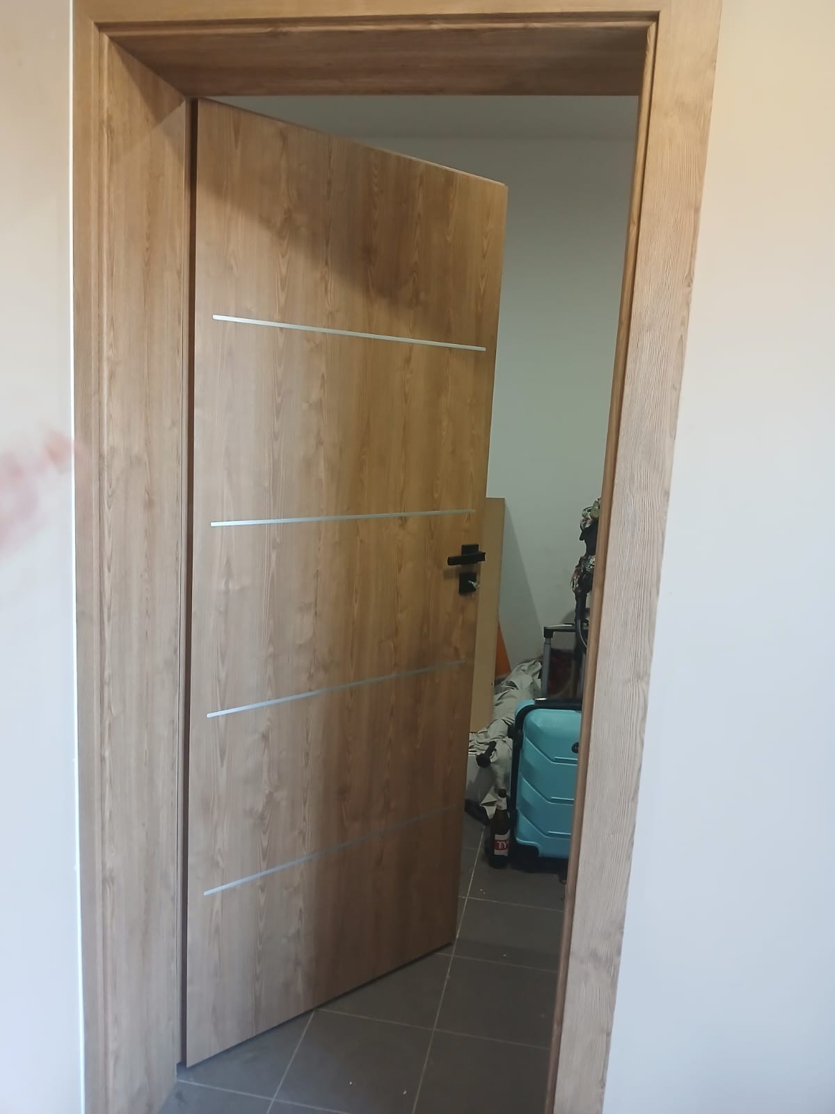 Otwarta, drewnopodobna wewnętrzna drzwi z poziomymi, srebrnymi paskami i czarną klamką, widok z boku na szarą podłogę i fragment pokoju z walizką.