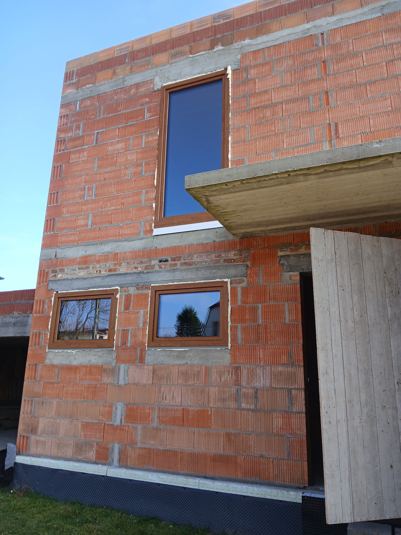 Surowy mur z brązowymi oknami PCV w trakcie budowy domu, widoczne pustaki ceramiczne i betonowe nadproża. Częściowo otwarte, drewniane drzwi wejściowe.