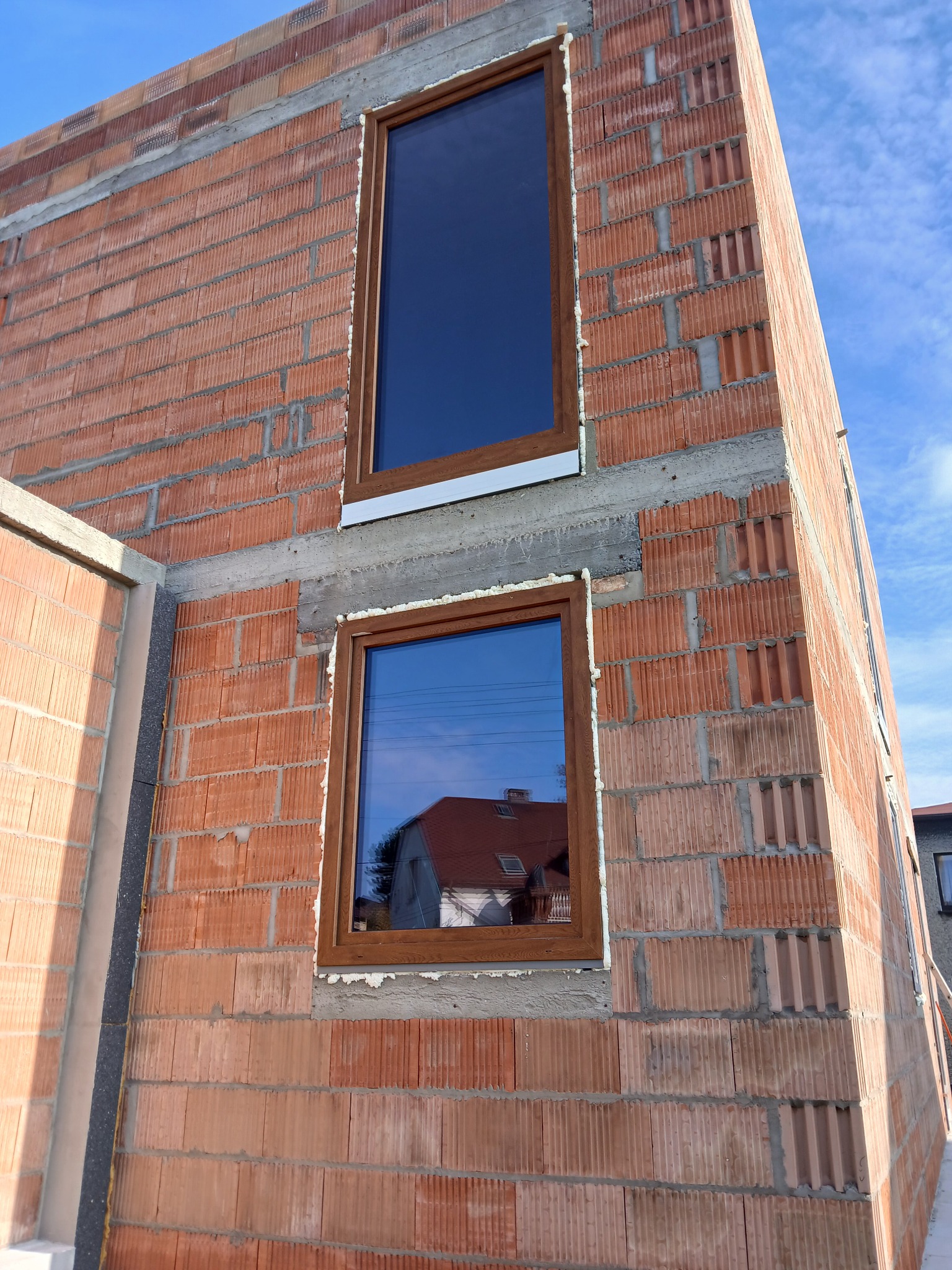 Montaż okien z brązową ramą w surowym murze z pustaków ceramicznych. Widoczne pianowanie i odbicie budynku w szybie. Błękitne niebo w tle.