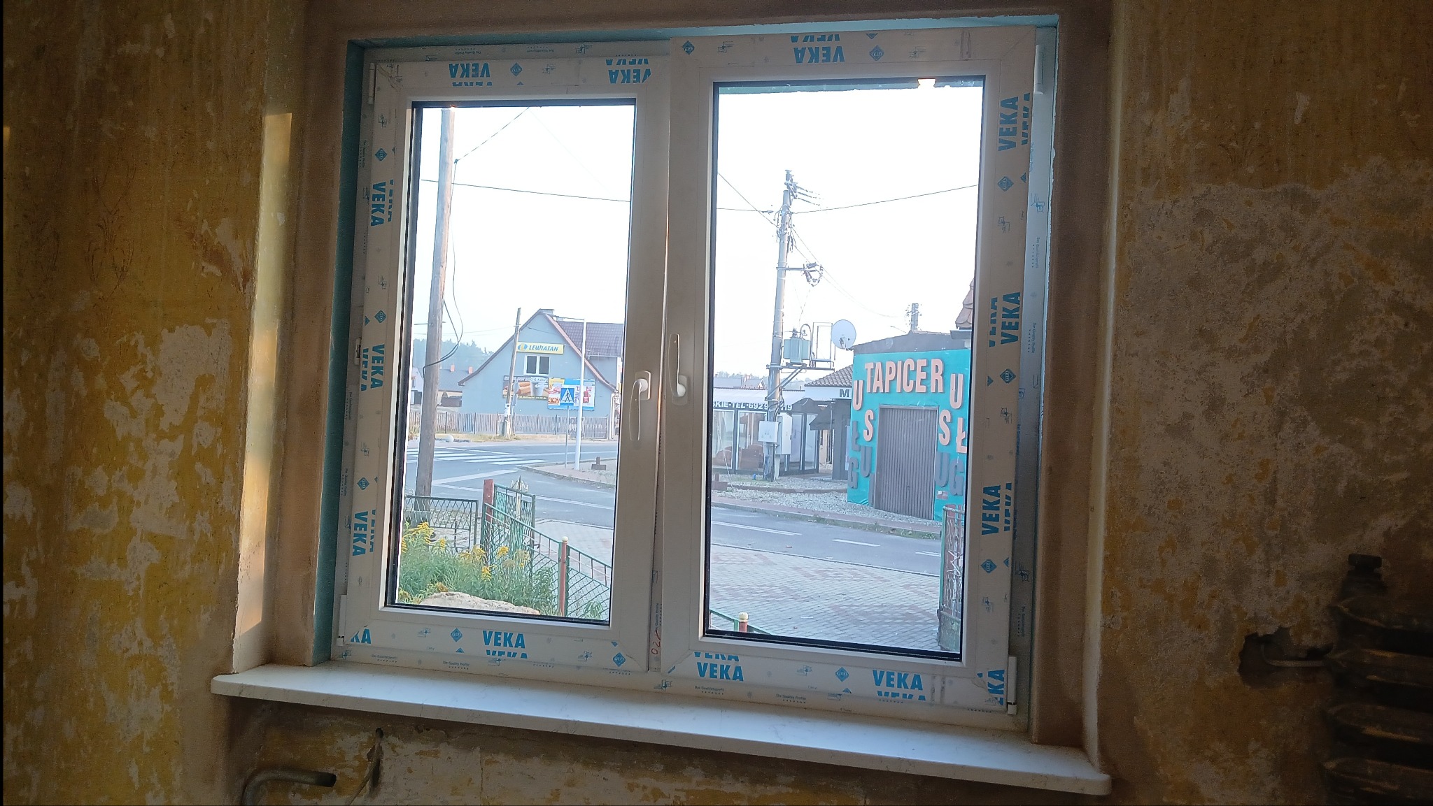Białe okno PCV marki VEKA, widok na ulicę z budynkami i sklepem 'Tapicer', ściany w surowym stanie przed wykończeniem. Wnętrze w trakcie remontu.