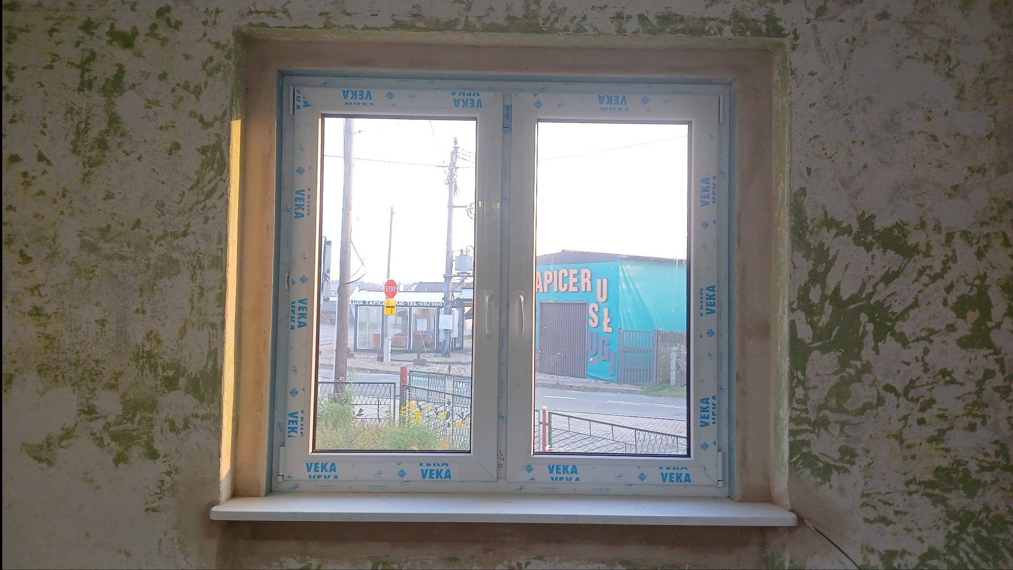 Białe okno PCV marki VEKA w surowym otoczeniu, z widokiem na ulicę i budynki. Ściany w zielonkawej fakturze, surowy mur wokół okna. Dziennie światło z zewnątrz.