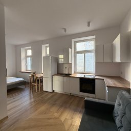 Wis-Bud Jakub Wistehube - Jasne, przestronne wnętrze mieszkania po remoncie, z widoczną kuchnią w zabudowie, drewnianym blatem, stołem z krzesłami oraz częścią sypialną z łóżkiem, oświetlone naturalnym światłem z okien.