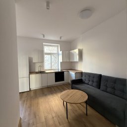 Wis-Bud Jakub Wistehube - Jasne wnętrze po remoncie: drewniana podłoga, białe meble kuchenne, szara sofa i okrągły stolik kawowy z czarnymi nogami.