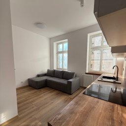 Wis-Bud Jakub Wistehube - Jasne wnętrze mieszkania po remoncie: szara sofa narożna z weluru, drewniana podłoga, zabudowa kuchenna z blatem imitującym drewno, okna z widokiem na budynek.