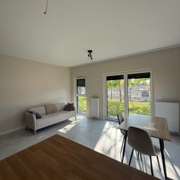 Wis-Bud Jakub Wistehube - Jasne wnętrze z beżowymi ścianami, sofa i stół z krzesłami w świetle słonecznym wpadającym przez okna balkonowe. Widoczne niedokończone podłączenie lampy sufitowej.