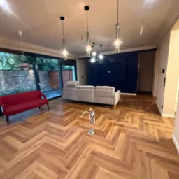 Salon z parkietem w jodełkę, szarą sofą i czerwoną kanapą. Nowoczesne lampy wiszące. Widok na ogród przez przeszklone drzwi.