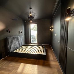 Wis-Bud Jakub Wistehube - Ciemne wnętrze sypialni po metamorfozie: parkiet w jodełkę, czarne ściany z dekoracyjnymi panelami, łóżko z szarym zagłówkiem i designerskie lampy.