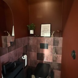 Wis-Bud Jakub Wistehube - Stylowa, mała łazienka w odcieniach bordo i czerni. Umywalka z szafką, wisząca toaleta, płytki na ścianach i podłodze w szachownicę. Dekoracje: roślina, plakat.