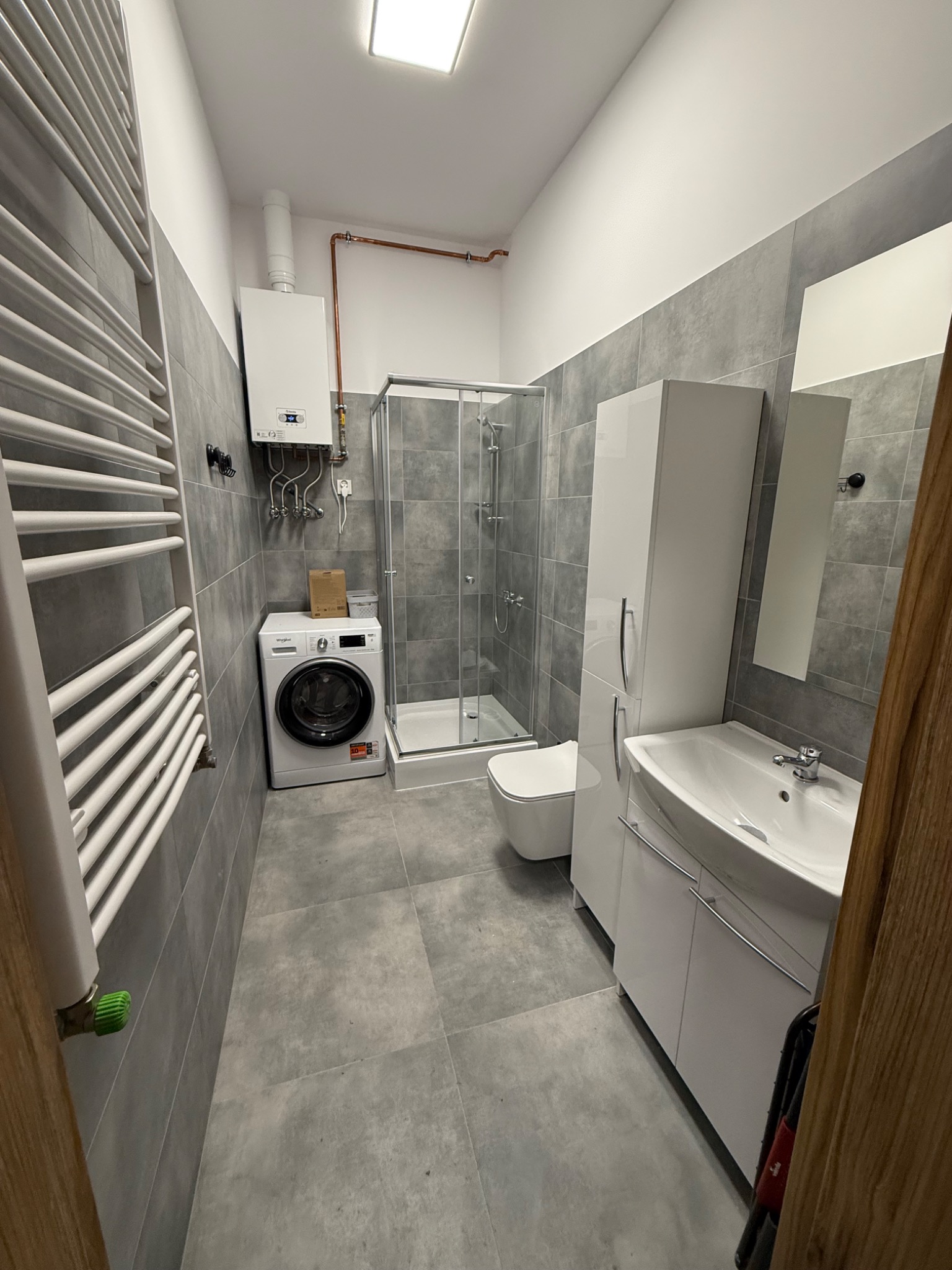 Nowoczesna łazienka w odcieniach szarości z pralką, kabiną prysznicową i białymi meblami łazienkowymi, widoczny grzejnik drabinkowy z zielonym zaworem.