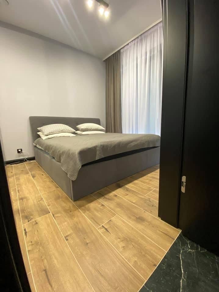 Nowoczesna sypialnia z drewnopodobną podłogą, szarą tapicerowaną ramą łóżka i białymi zasłonami w tle, widok z perspektywy wejścia.