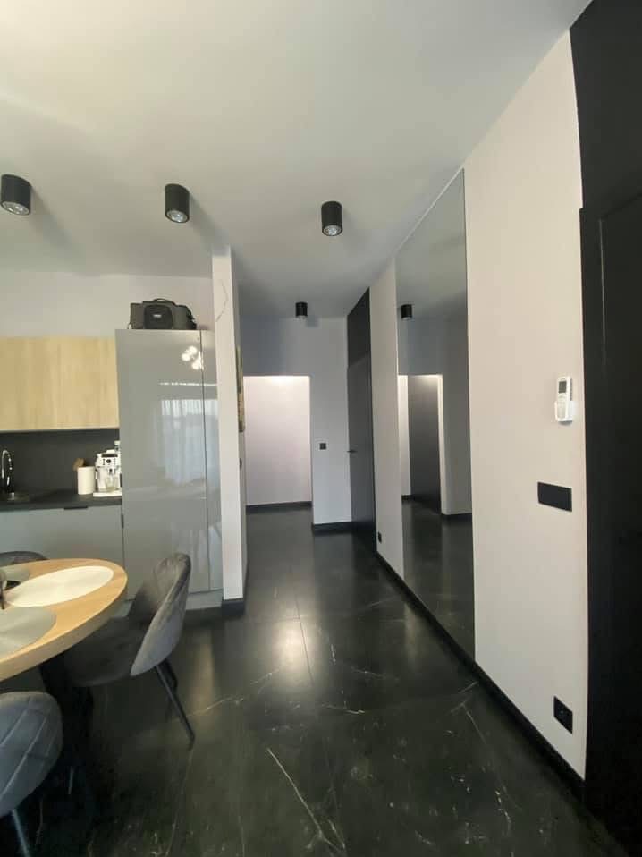 Nowoczesne wnętrze mieszkania z czarną marmurową podłogą, jasnymi ścianami, czarnymi akcentami i dużym lustrem, widok na kuchnię i korytarz.