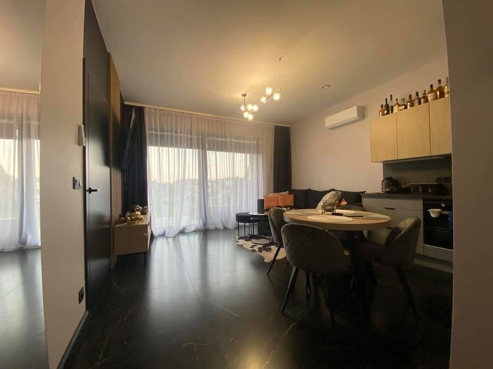 Nowoczesne wnętrze mieszkania z czarną, błyszczącą podłogą, okrągłym stołem jadalnym z szarymi krzesłami, kuchnią w tle i dużym oknem z firanami wpadającymi w biel.