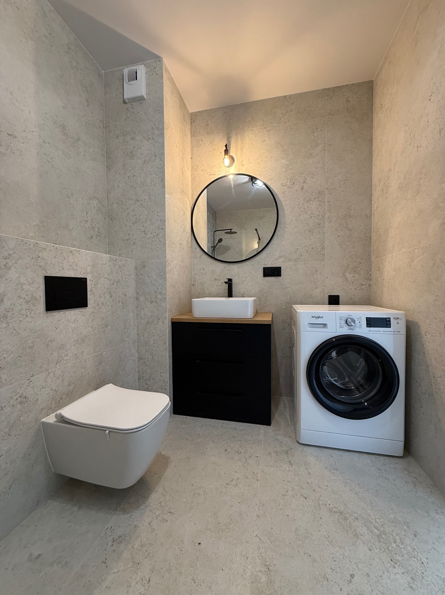 Nowoczesna łazienka z płytkami imitującymi kamień, wiszącą toaletą z czarnym przyciskiem, czarną szafką z drewnianym blatem, okrągłym lustrem z oświetleniem oraz białą pralką.