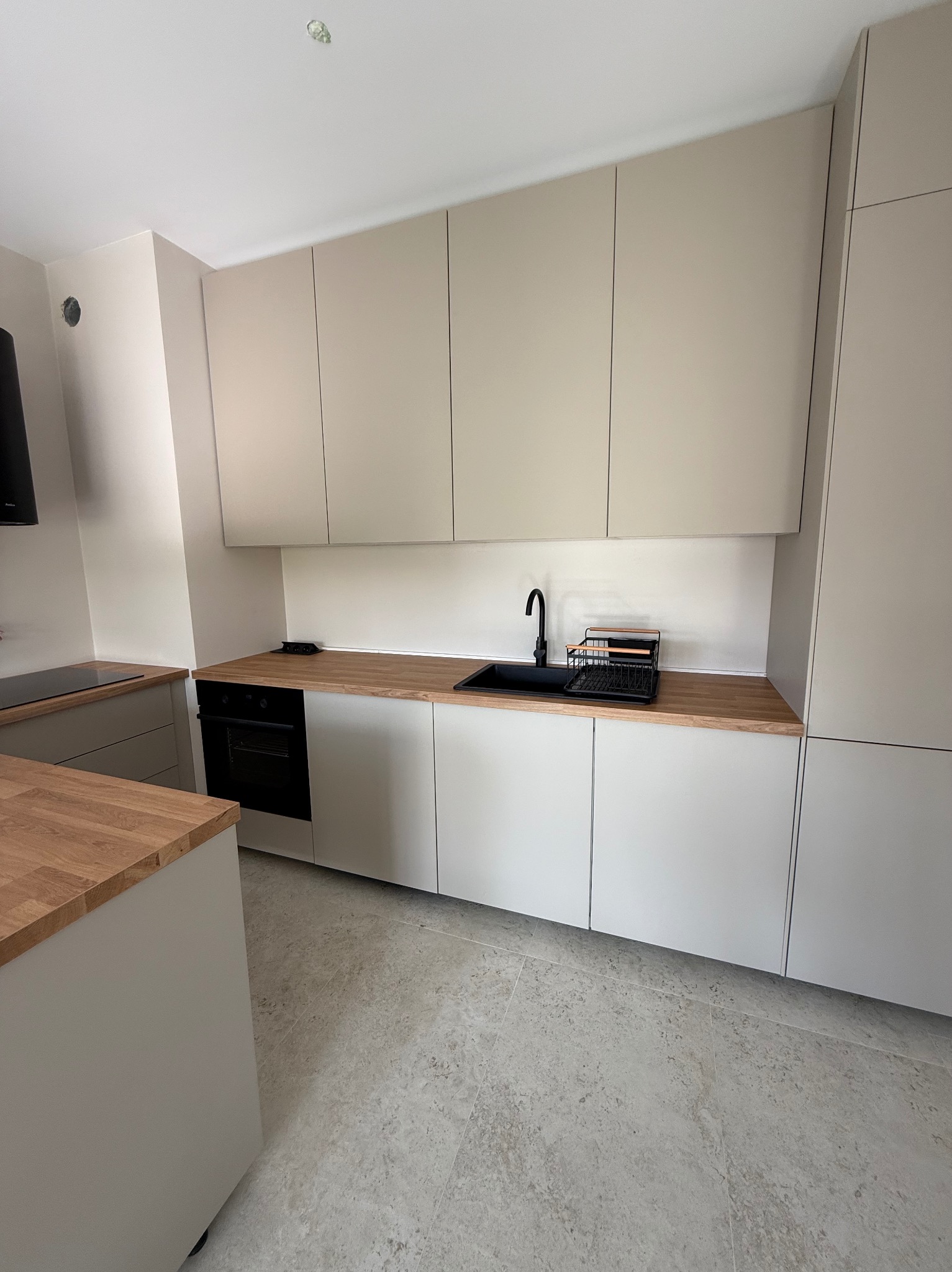 Minimalistyczna kuchnia w odcieniach szarości z drewnianym blatem i czarną armaturą. Górne szafki bez uchwytów, na podłodze szare płytki imitujące kamień.