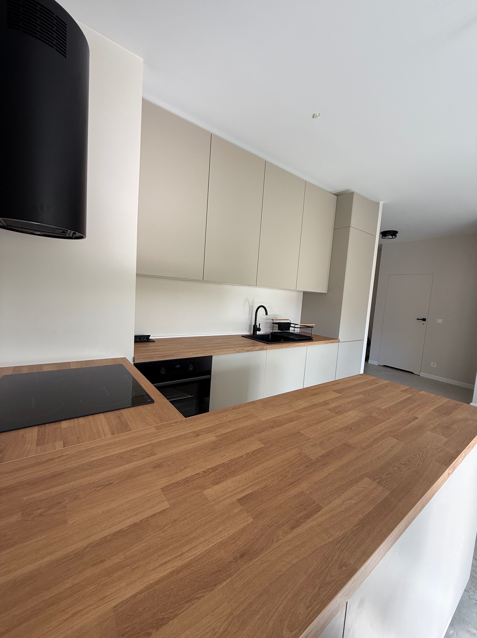 Minimalistyczna kuchnia z drewnianym blatem, czarną armaturą i płytą indukcyjną. Jasne szafki wiszące i neutralne ściany tworzą spójną całość.