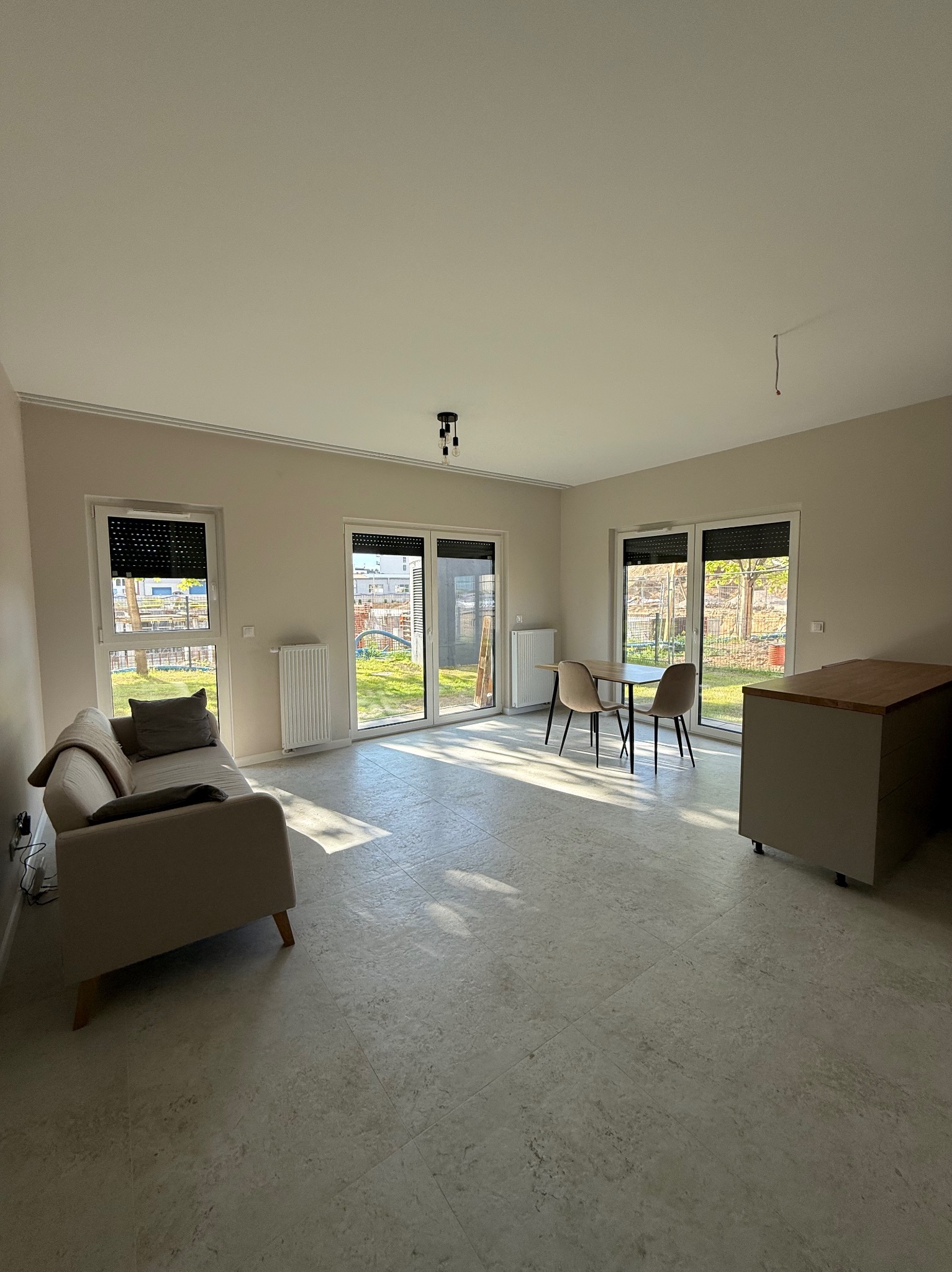 Jasne wnętrze po remoncie: sofa, stół z krzesłami, okna z roletami. Widoczne grzejniki i fragment zabudowy kuchennej. Na podłodze jasne płytki imitujące kamień.