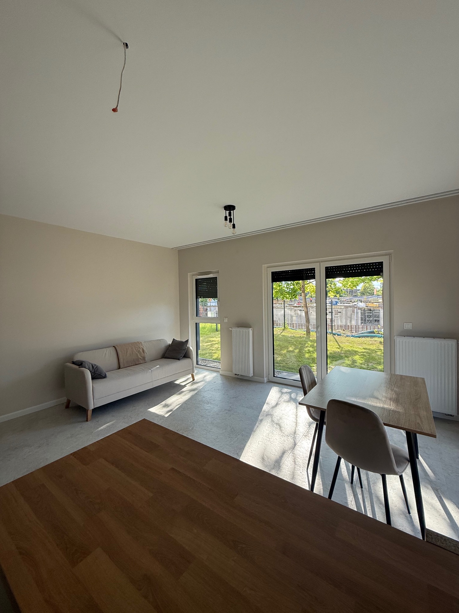 Jasne wnętrze z beżowymi ścianami, sofa i stół z krzesłami w świetle słonecznym wpadającym przez okna balkonowe. Widoczne niedokończone podłączenie lampy sufitowej.