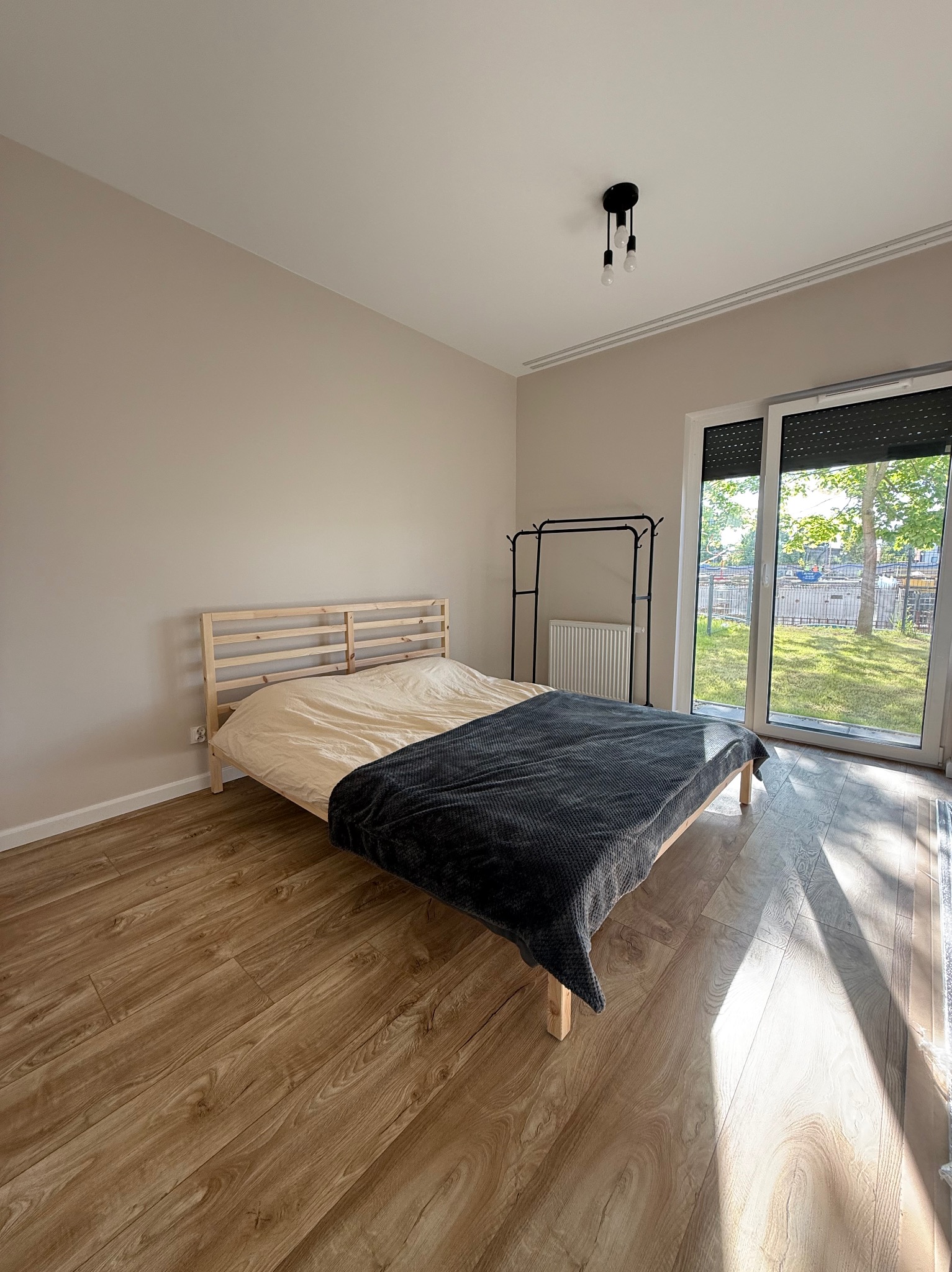 Jasne wnętrze sypialni po remoncie: drewniana podłoga, łóżko przy oknie z widokiem na zieleń. Minimalistyczny wystrój, ciepłe oświetlenie.