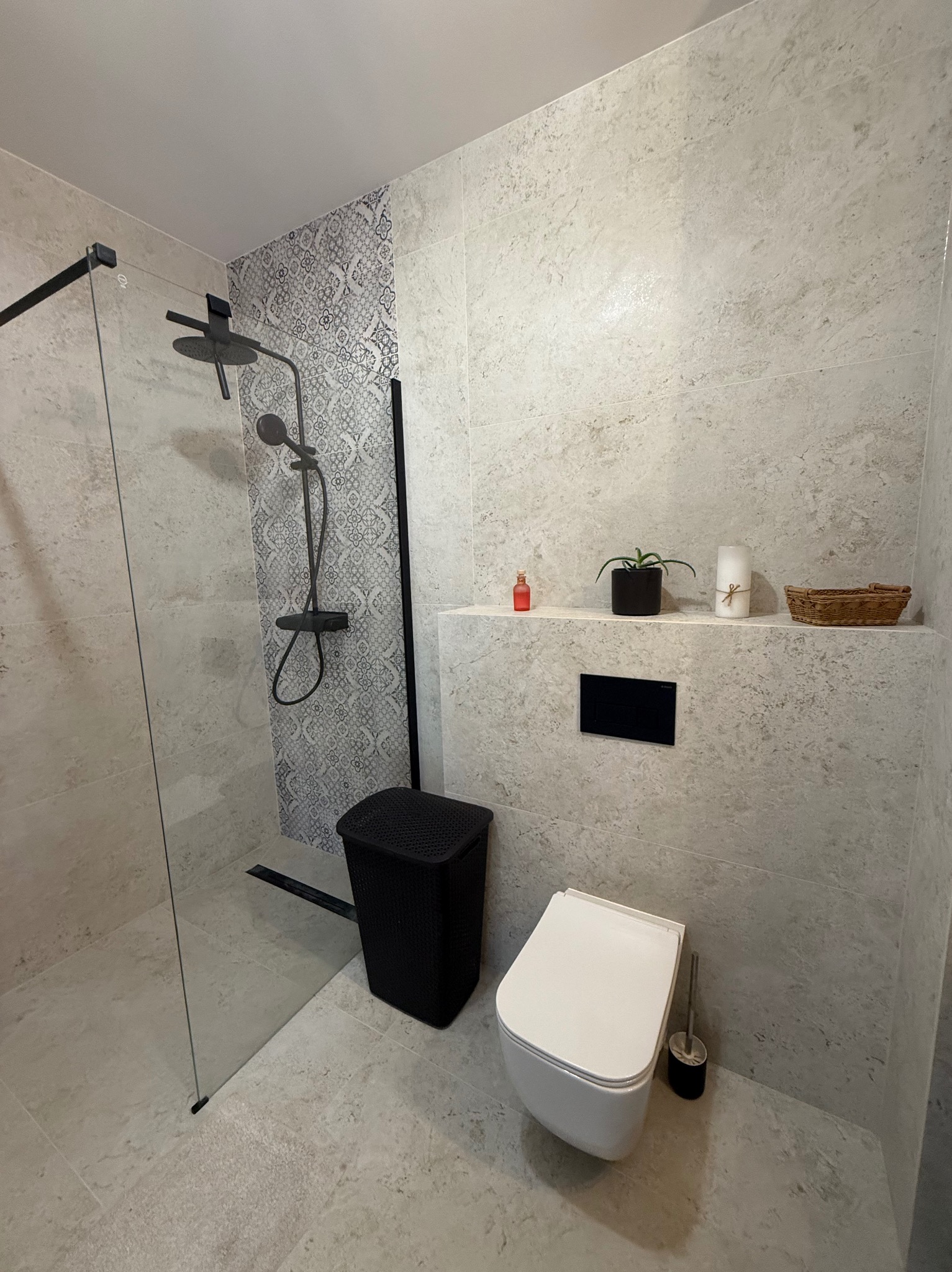 Nowoczesna łazienka z kabiną typu walk-in, szarą armaturą i płytkami imitującymi kamień. Uwagę zwraca dekoracyjna ściana z wzorzystymi płytkami nad strefą prysznica.