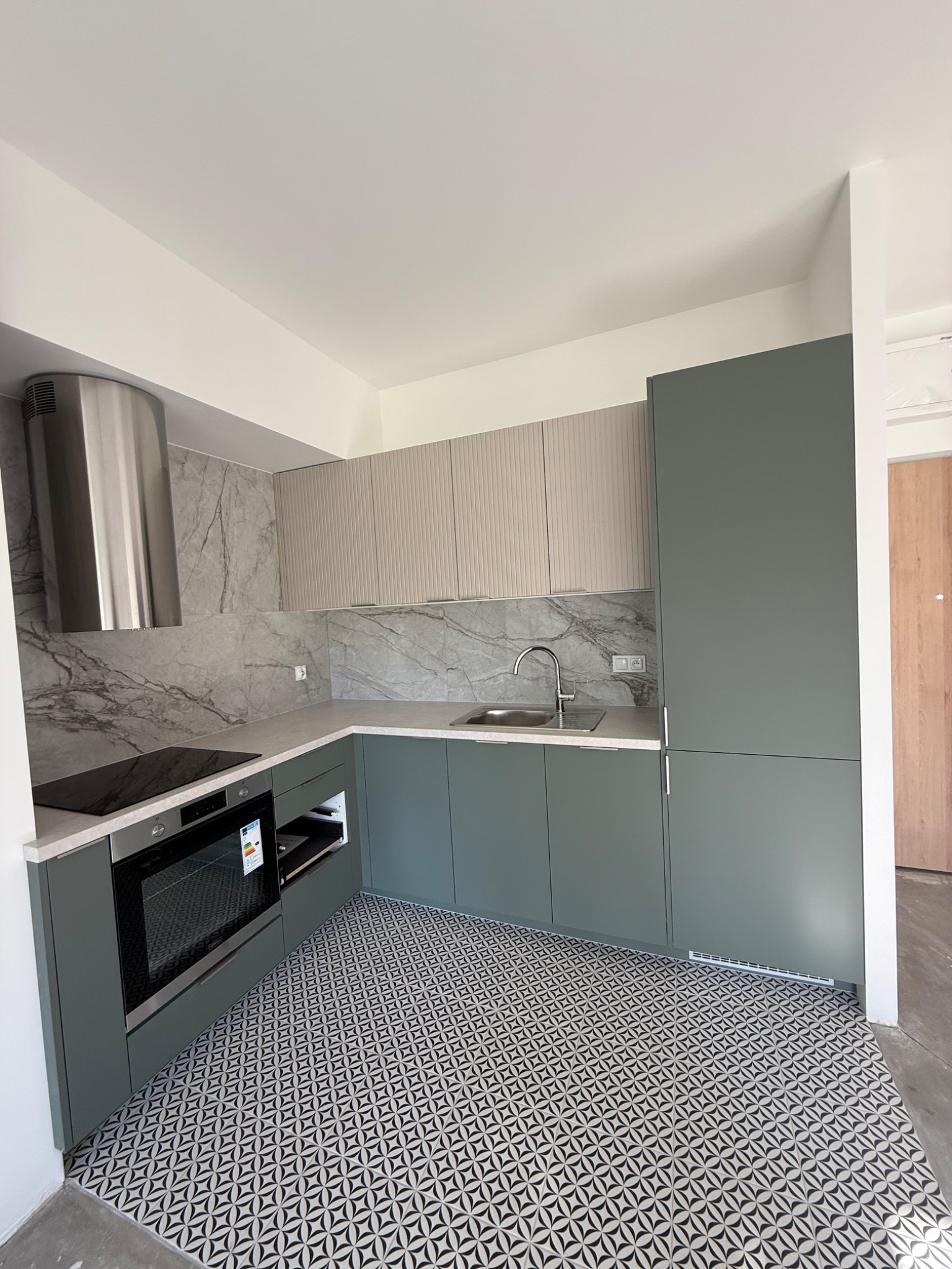 Nowoczesna kuchnia w odcieniach szarości i zieleni z wzorzystą podłogą. Widok na zabudowę kuchenną, okap i płytki imitujące marmur na ścianie.
