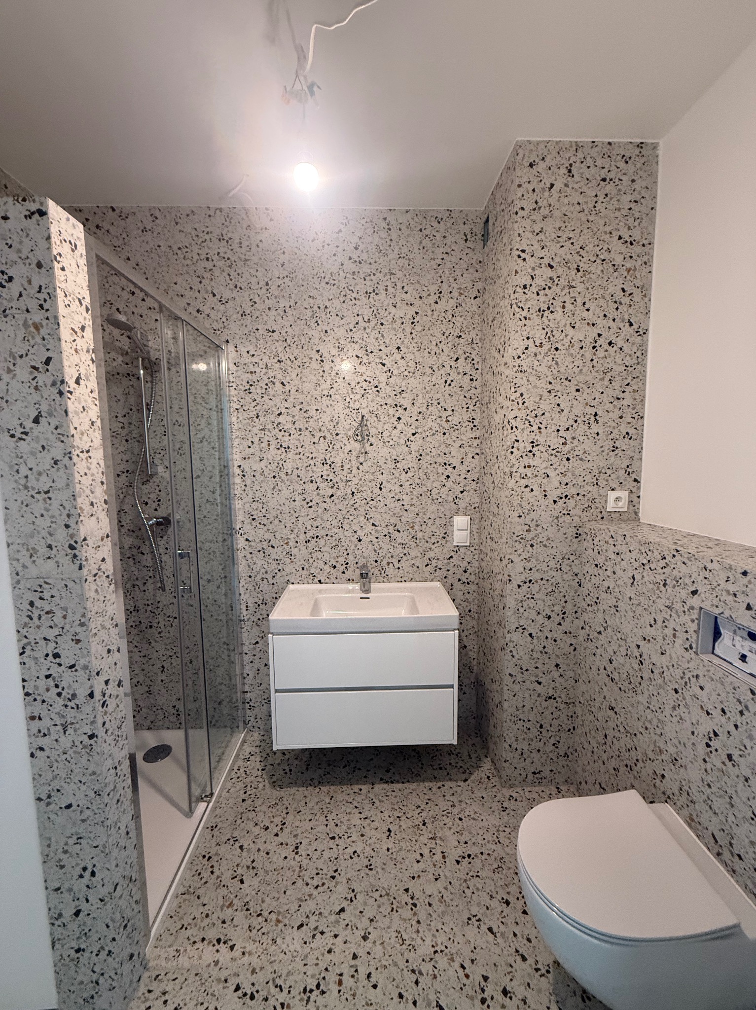 Wykończona łazienka z kabiną prysznicową, umywalką z szafką i toaletą. Ściany i podłoga wyłożone płytkami imitującymi lastryko w odcieniach szarości i bieli. Oświetlenie sufitowe.