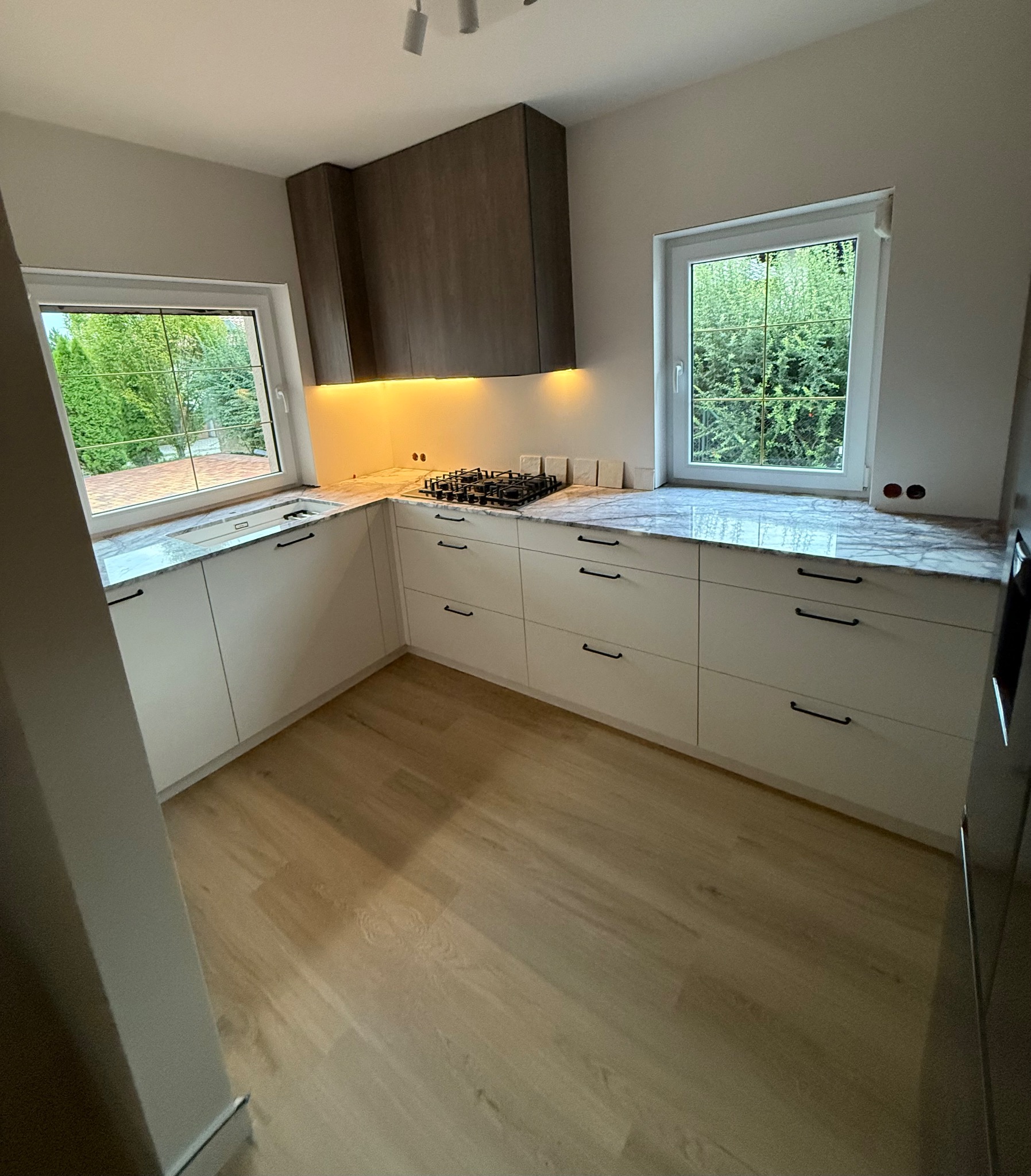 Nowoczesna kuchnia z białymi szafkami i marmurowym blatem pod oknem z widokiem na zieleń. Drewniane szafki wiszące z oświetleniem LED. Jasna drewniana podłoga.