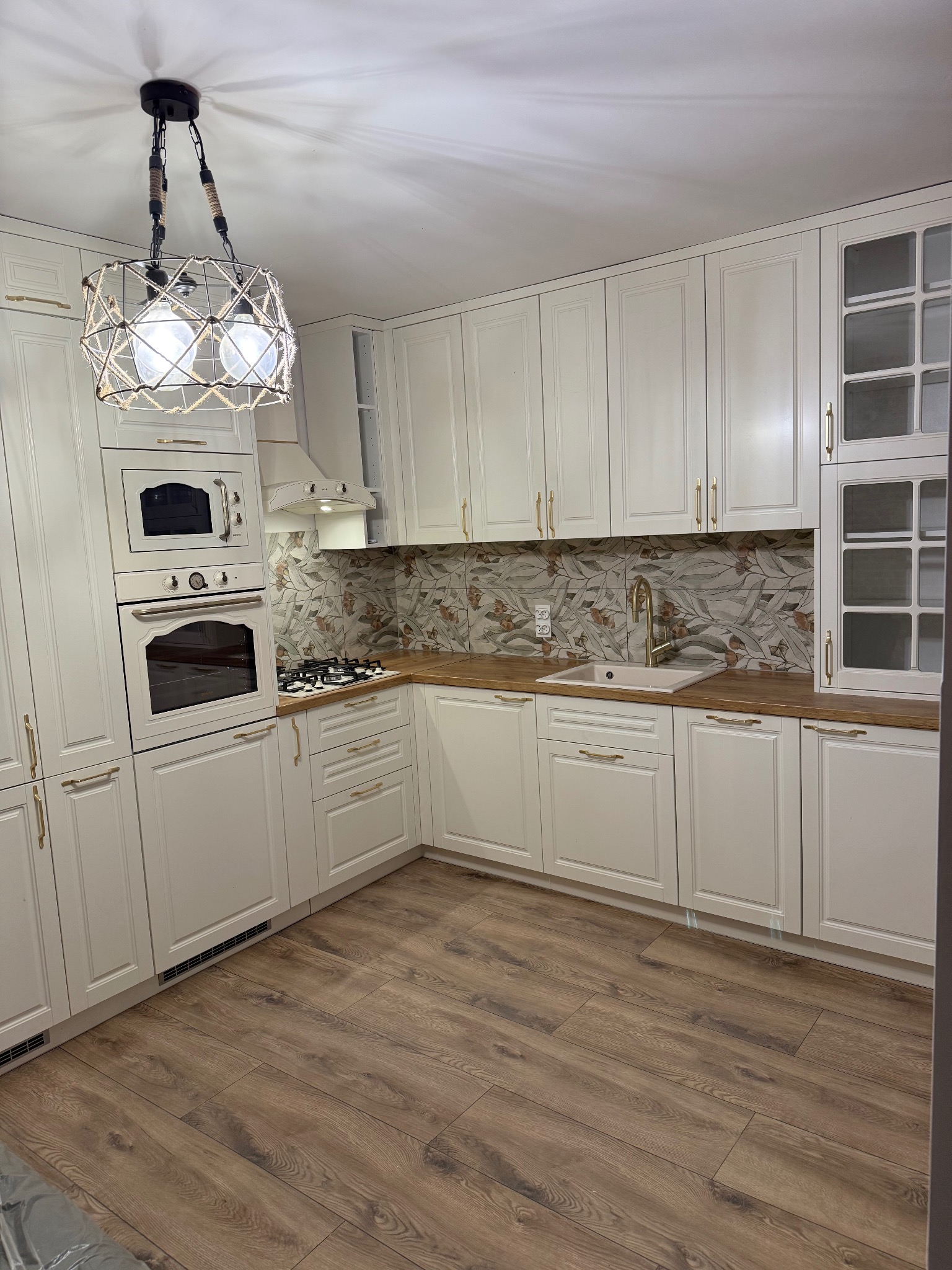 Jasna kuchnia w stylu klasycznym: kremowe szafki z frezowanymi frontami, drewniany blat, dekoracyjna lampa z linami, płytki z motywem roślinnym nad blatem, drewniana podłoga.