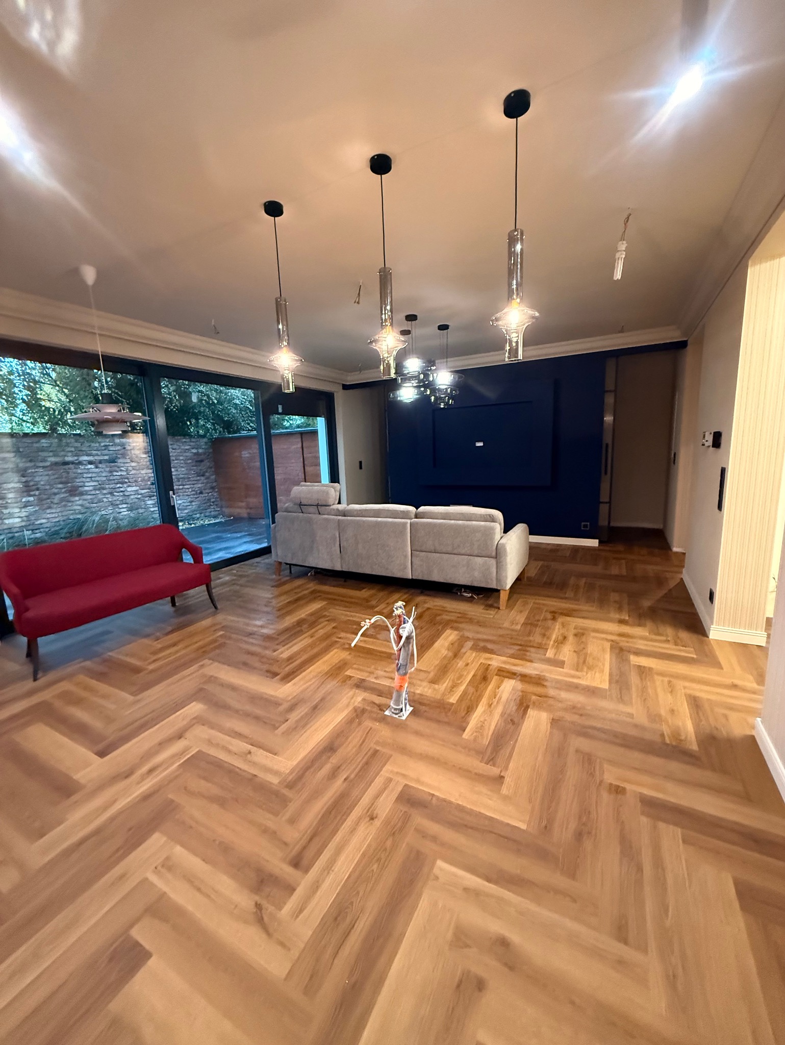 Salon z parkietem w jodełkę, szarą sofą i czerwoną kanapą. Nowoczesne lampy wiszące. Widok na ogród przez przeszklone drzwi.