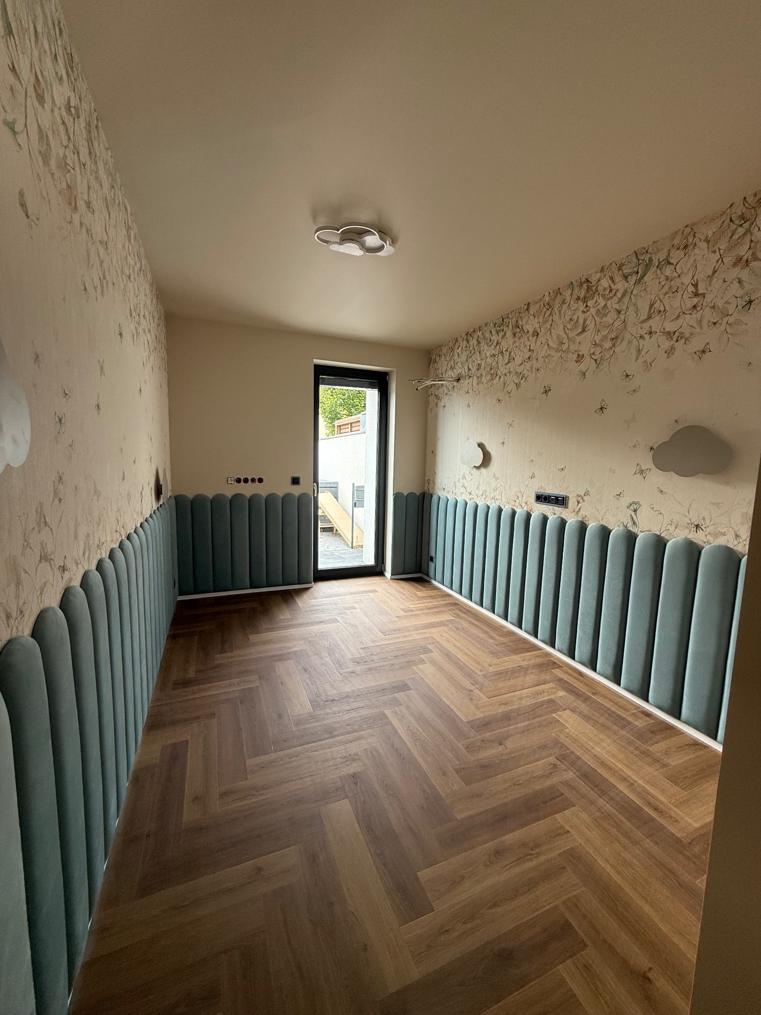 Pokój po wykończeniu: parkiet w jodełkę, tapeta w kwiaty, panele ścienne w kolorze morskim i dekoracyjne chmurki. Nowoczesne oświetlenie sufitowe.