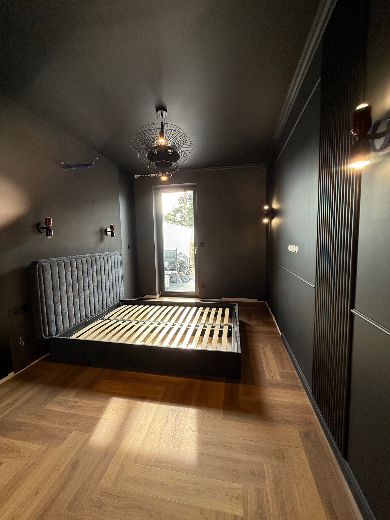 Ciemne wnętrze sypialni po metamorfozie: parkiet w jodełkę, czarne ściany z dekoracyjnymi panelami, łóżko z szarym zagłówkiem i designerskie lampy.