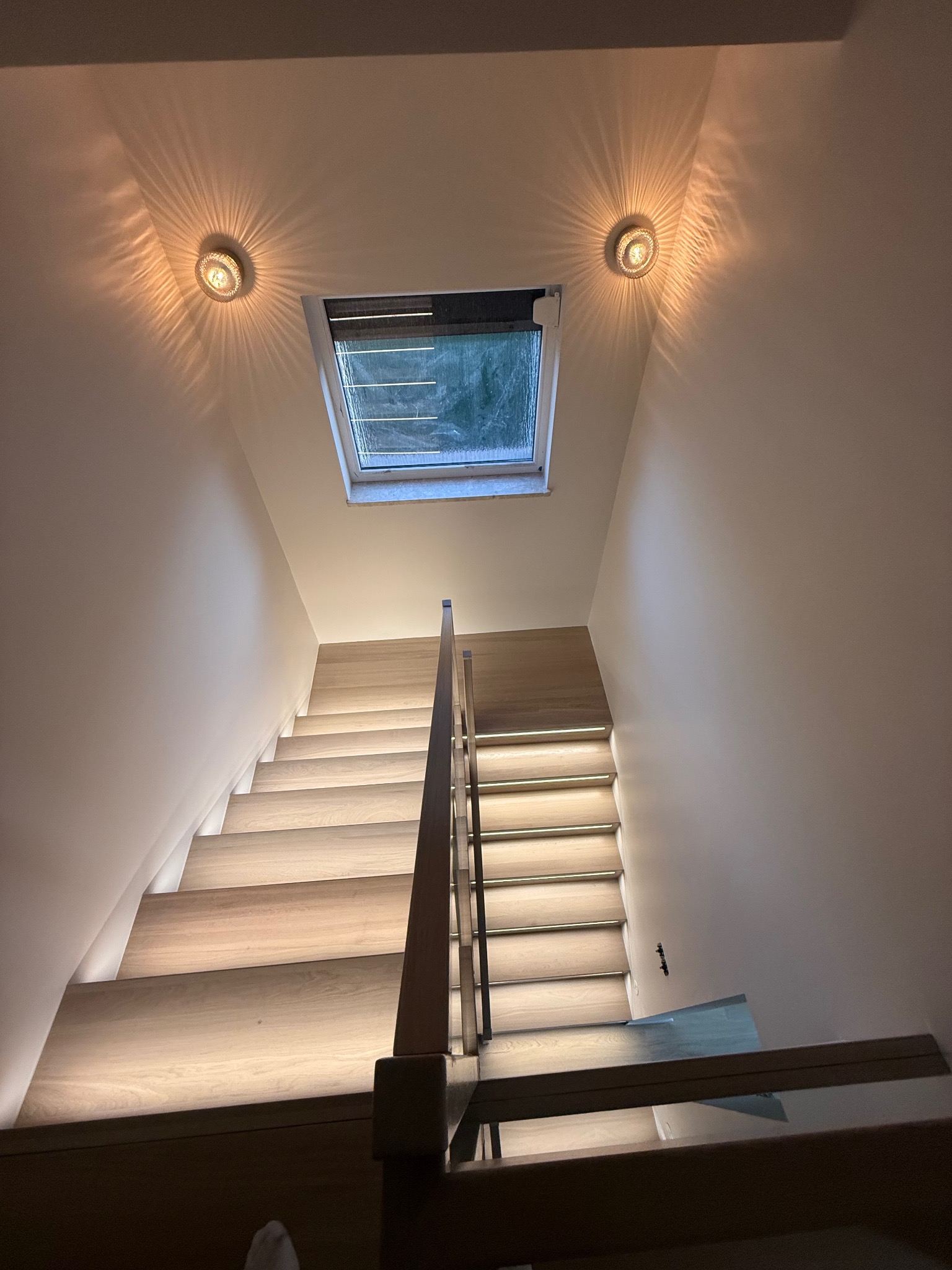 Nowoczesne schody z podświetleniem LED, drewniane stopnie, szklana balustrada, oświetlenie sufitowe punktowe, okno dachowe. Minimalistyczny design.