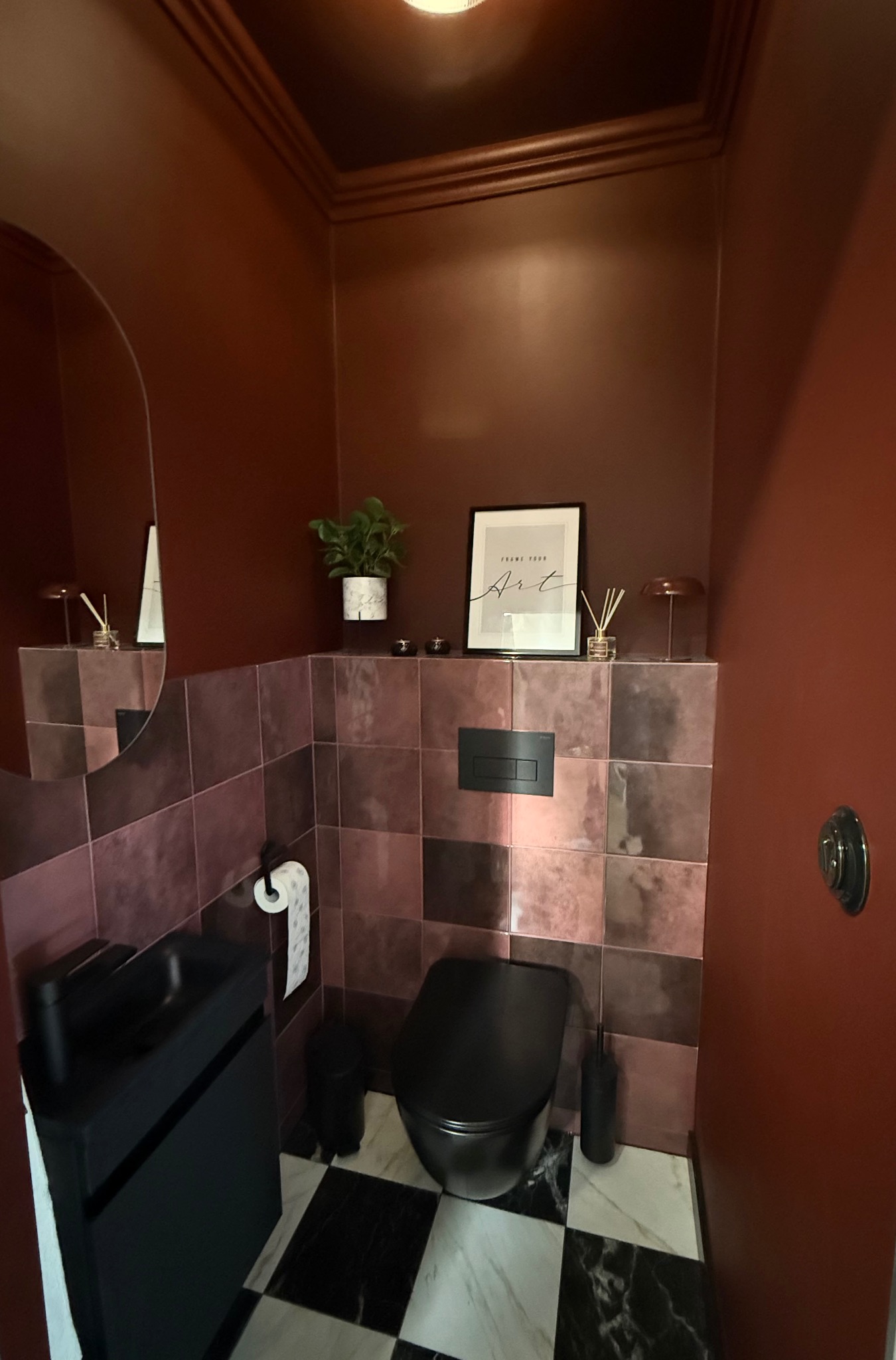 Stylowa, mała łazienka w odcieniach bordo i czerni. Umywalka z szafką, wisząca toaleta, płytki na ścianach i podłodze w szachownicę. Dekoracje: roślina, plakat.