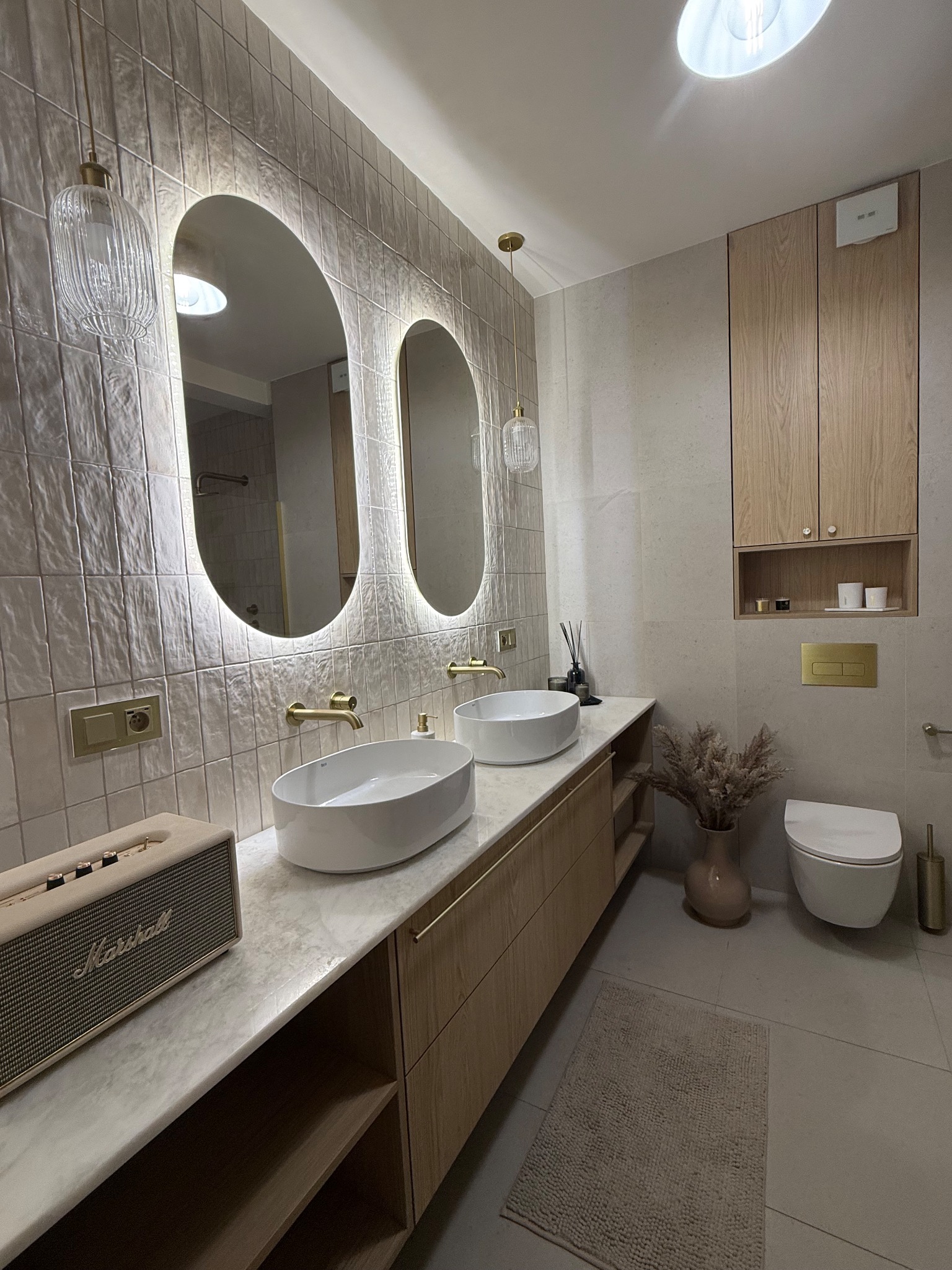 Nowoczesna łazienka z owalnymi lustrami LED, beżowymi płytkami i drewnianą szafką pod umywalką. Uwagę zwraca głośnik Marshall na blacie.