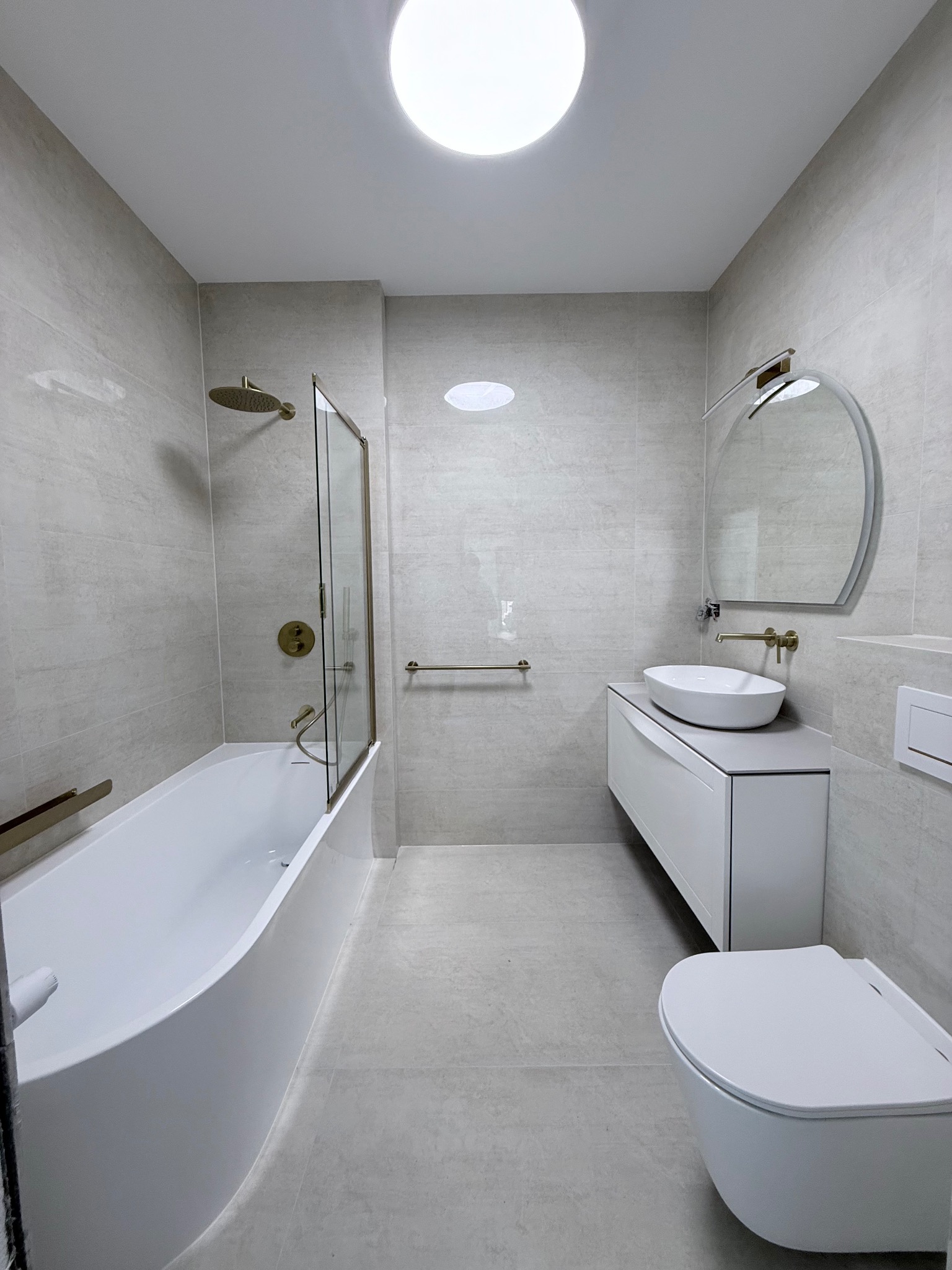 Jasna łazienka po remoncie: wanna z parawanem, umywalka na szafce, lustro owalne, toaleta podwieszana. Armatura w kolorze złotym. Płytki w odcieniach szarości.