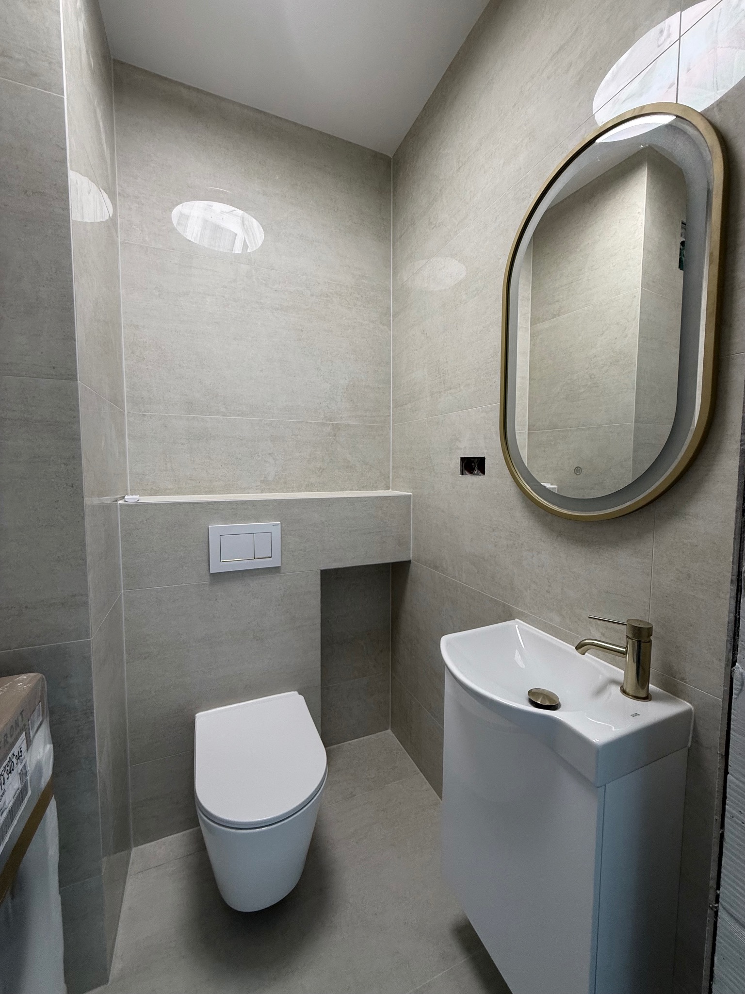 Nowoczesna łazienka w beżowych płytkach z owalnym lustrem w złotej ramie, wiszącą toaletą i umywalką z szafką. Minimalistyczny design, widoczne detale wykończenia.