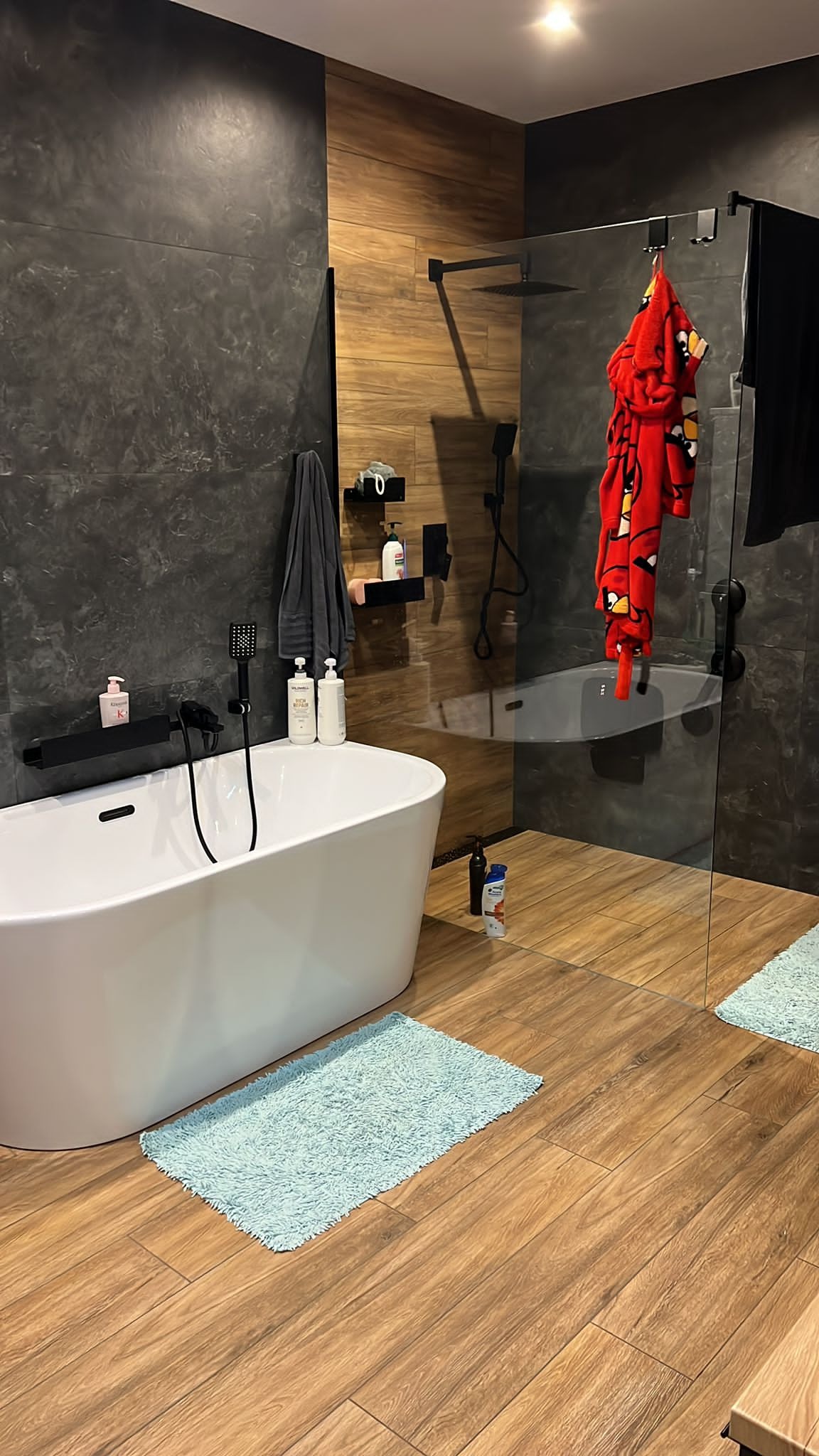 Nowoczesna łazienka z wolnostojącą białą wanną, drewnopodobną podłogą i szaro-czarnymi ścianami, z kabiną prysznicową typu walk-in i akcentem w postaci czerwonego szlafroka z motywem Angry Birds.