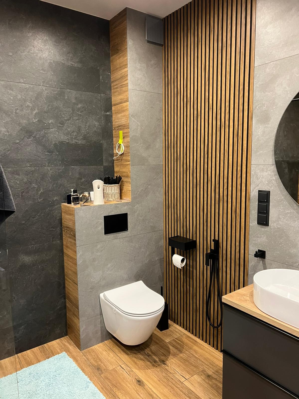 Nowoczesna łazienka z szarymi płytkami imitującymi kamień, drewnianymi lamelami na ścianie, podwieszaną toaletą, drewnianymi akcentami i błękitnym dywanikiem.